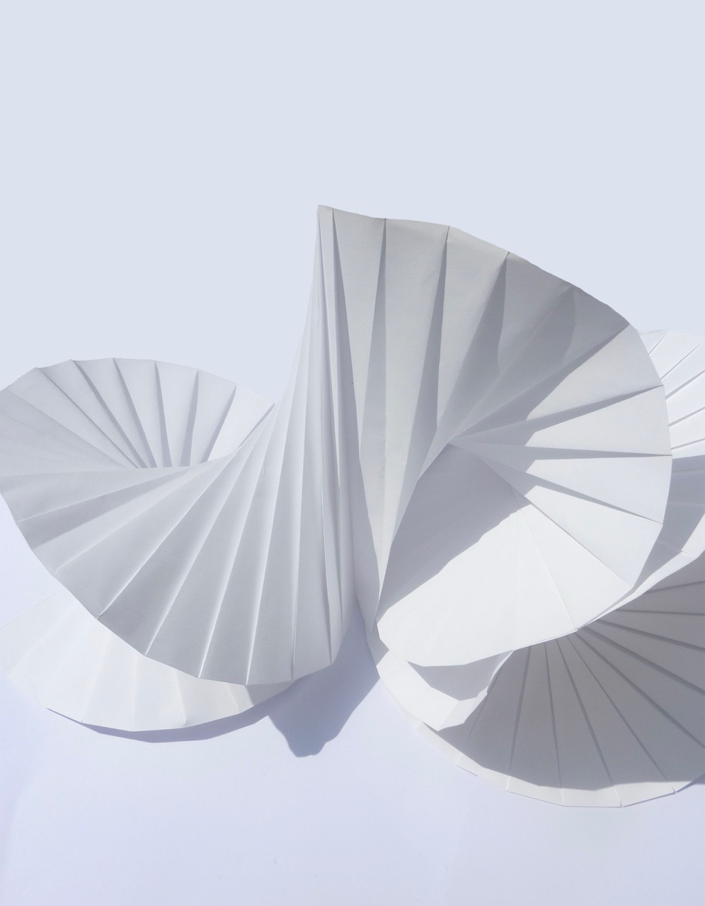 Abstract illustration with overlapping fan-shaped forms in soft pastels. Pleated semicircular shapes in pale pink, mint green, and lavender cast subtle shadows on light grey background.