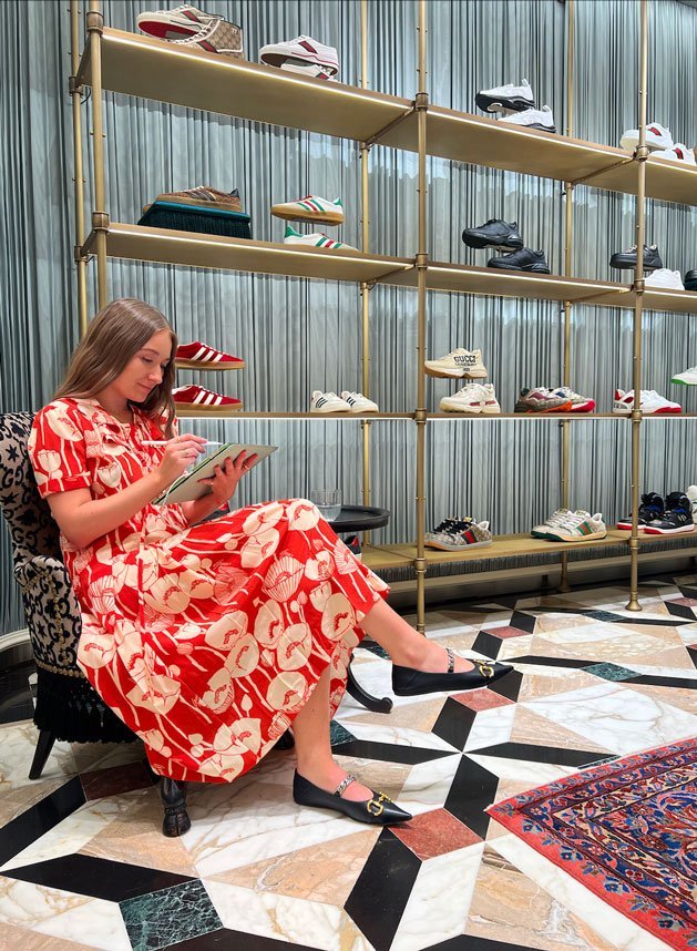 Tegan Price wearing a bright red dress, sat down drawing in front of racks of sneakers.