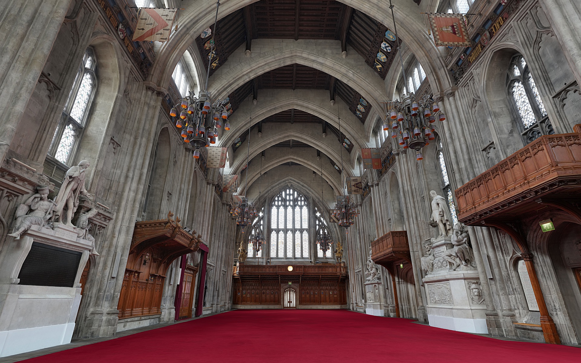 A vast Gothic church interior with a red carpeted floor, arched stone vaults and tall stained-glass windows, flanked by carved statues and wooden balconies.
