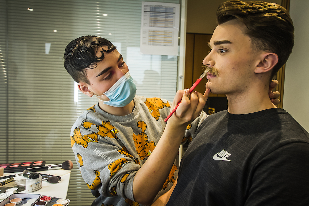 A makeup artist wearing a face mask applies a false moustache to an actor.