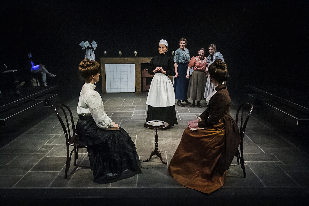 A performer dressed as a maid approaches two other performers in period costume sitting on chairs with a small table in between.