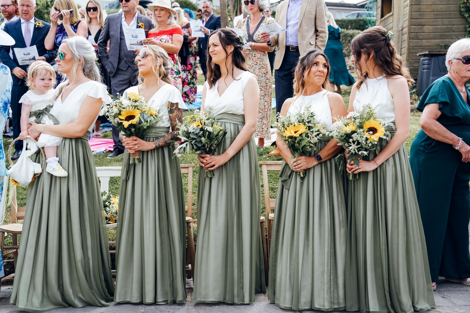 A line of five bridesmaids. Four are holding sunflowers, one is holding a child in her arms.