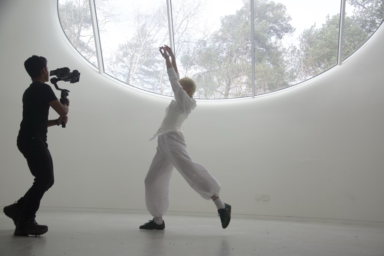 Person filming another person dancing in white room with large circular window showing trees outside.