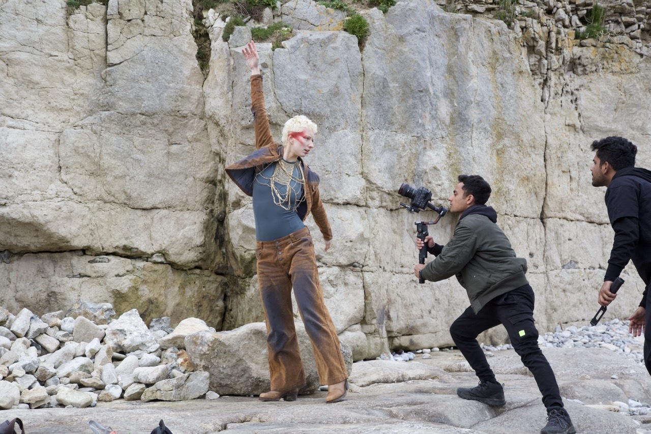 Film crew recording blonde woman in brown outfit against white chalk cliff face with rocky debris below.