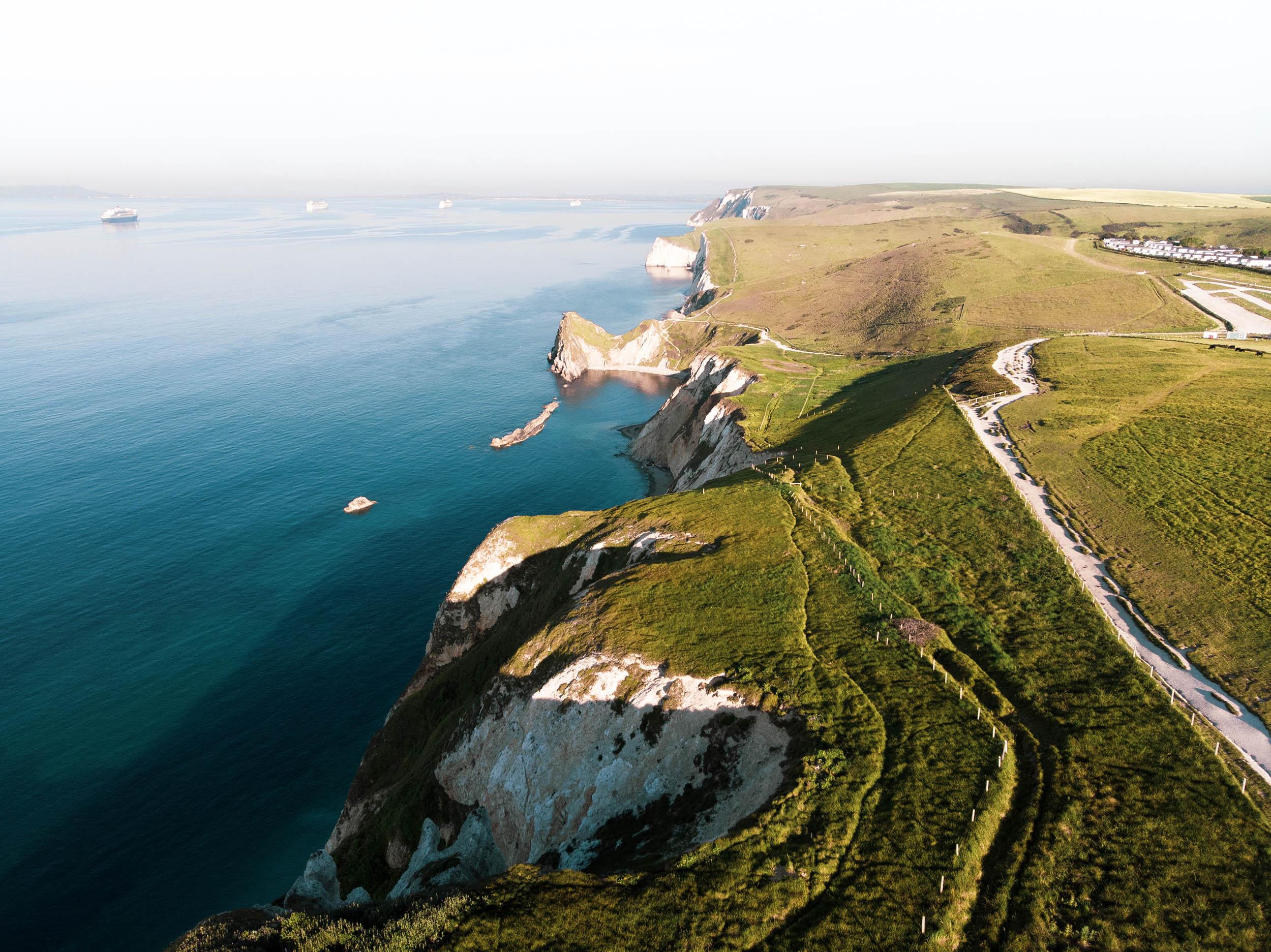Aerial photo showing the sea meeting clifftops, with greenery and pathways visible on the right.
