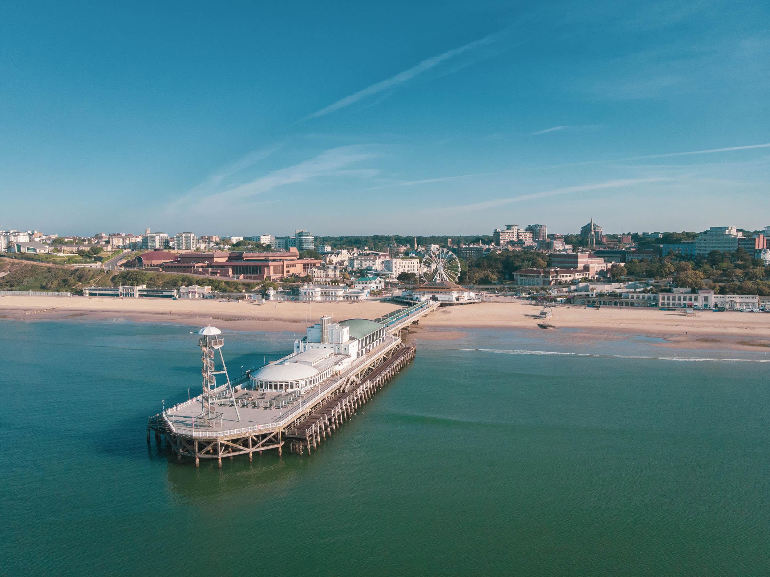 A pier in the middle of the sea leading back towards a beach with a townscape in the background.