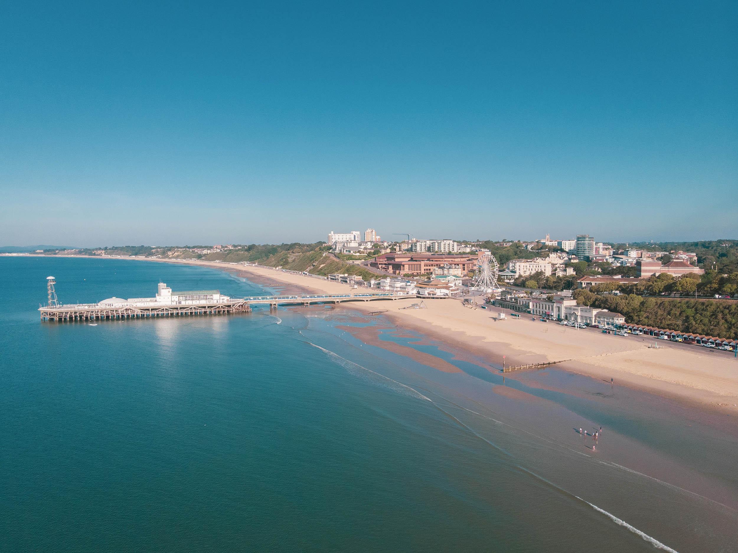 Aerial photo showing the sea, a long stretch of coastline and a pier coming from the beach into the water. A town with buildings is visible in the background.