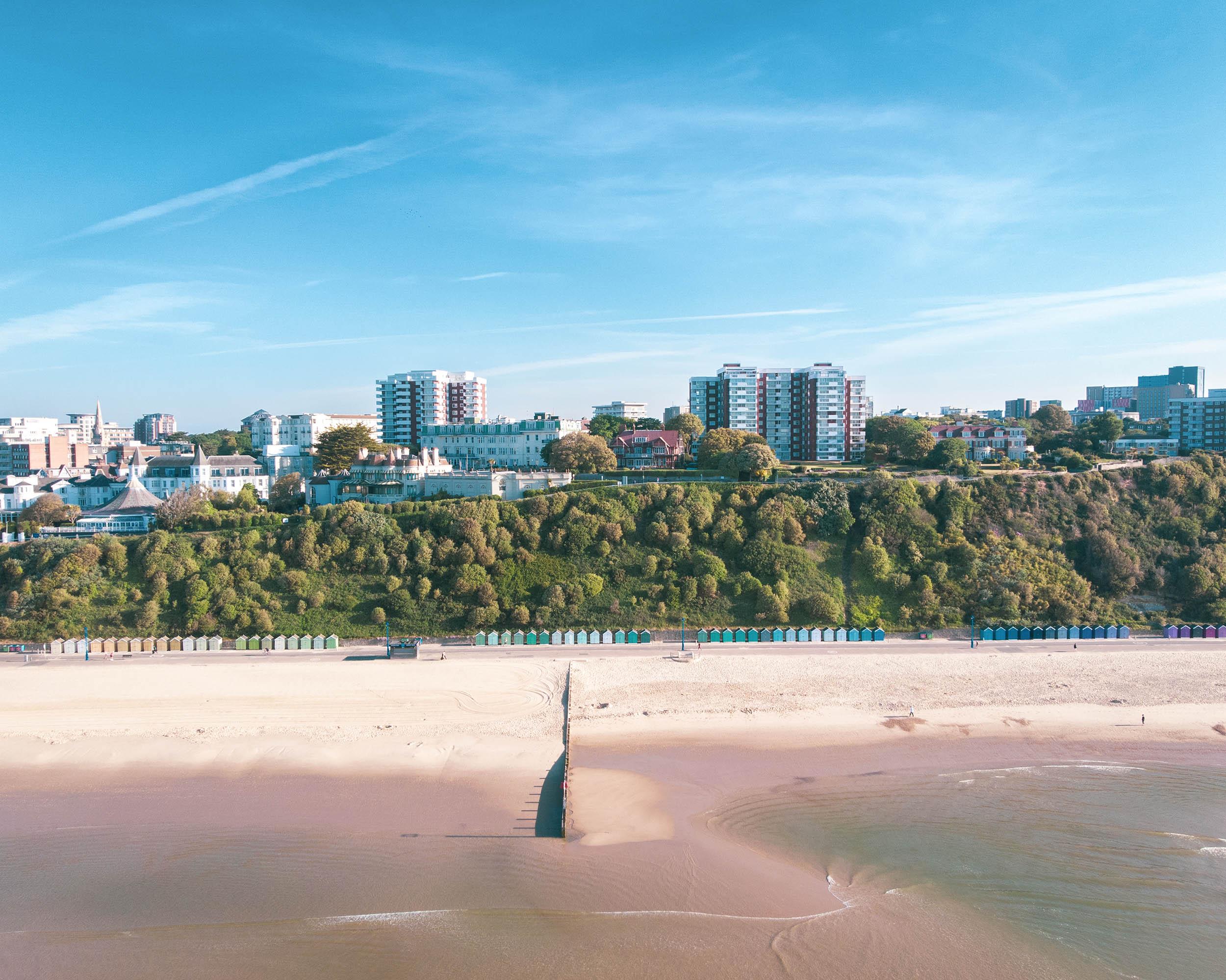 The sea meeting the sand of a beach, with rows of beach huts on the promenade, greenery behind them and a townscape in the background.
