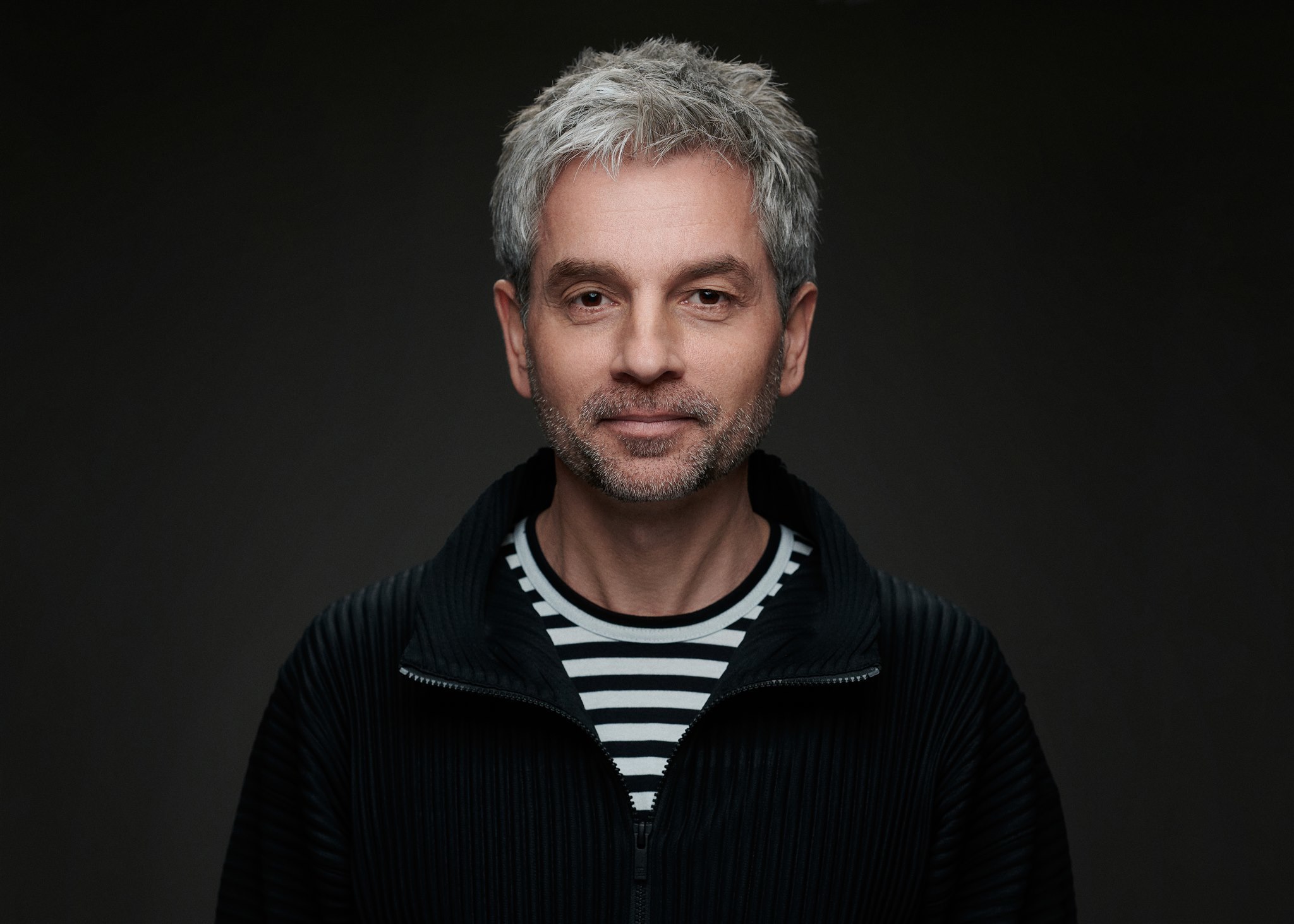 Man with grey hair and beard wearing black cardigan over striped shirt, smiling against dark background.