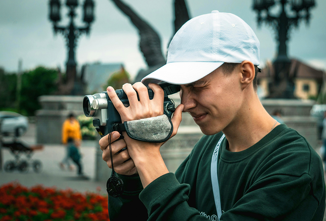 Close-up of a young person wearing a green top and white baseball cap peering into the viewfinder of a camcorder.
