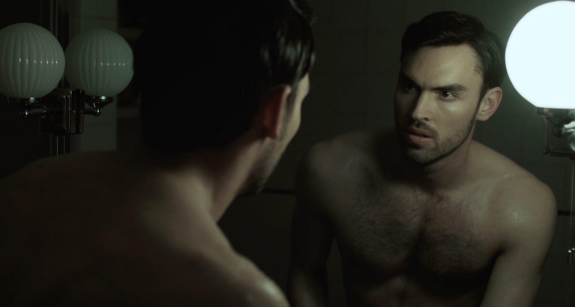 A shirtless man looks seriously at his reflection in a mirror.