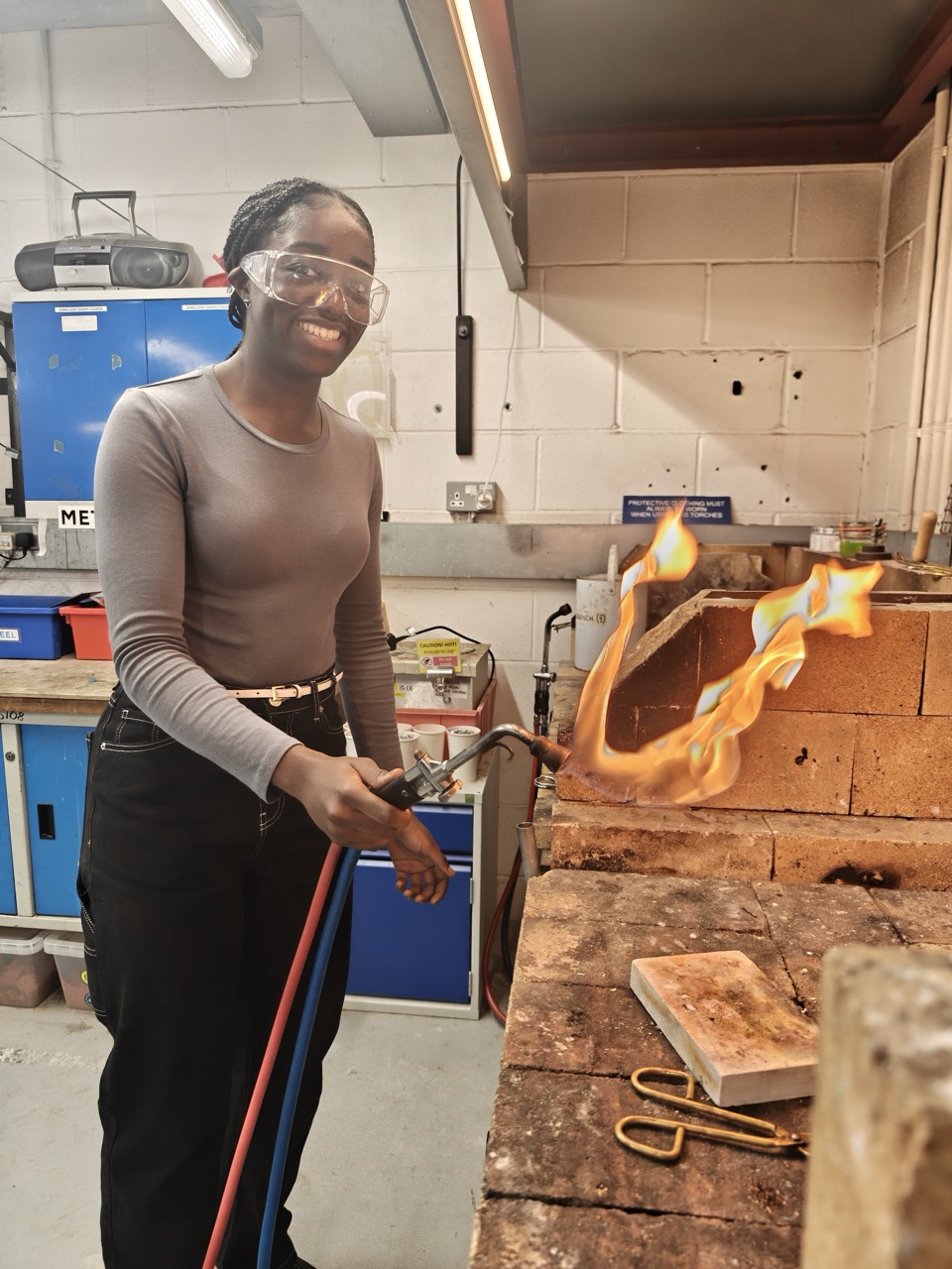 A woman wearing protective goggles and a long-sleeved grey top holds a blowtorch directed at a metal object, which is producing flames. She stands in a workshop with a workbench and blue storage cabinets in the background.