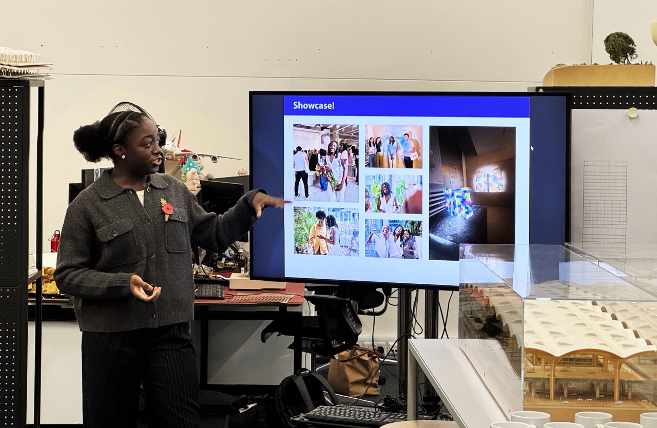 A woman in a grey jacket gestures towards a presentation screen showing a collage of colourful photographs and visual displays. The room features a model structure and a range of desk items, with natural light coming from the windows.
