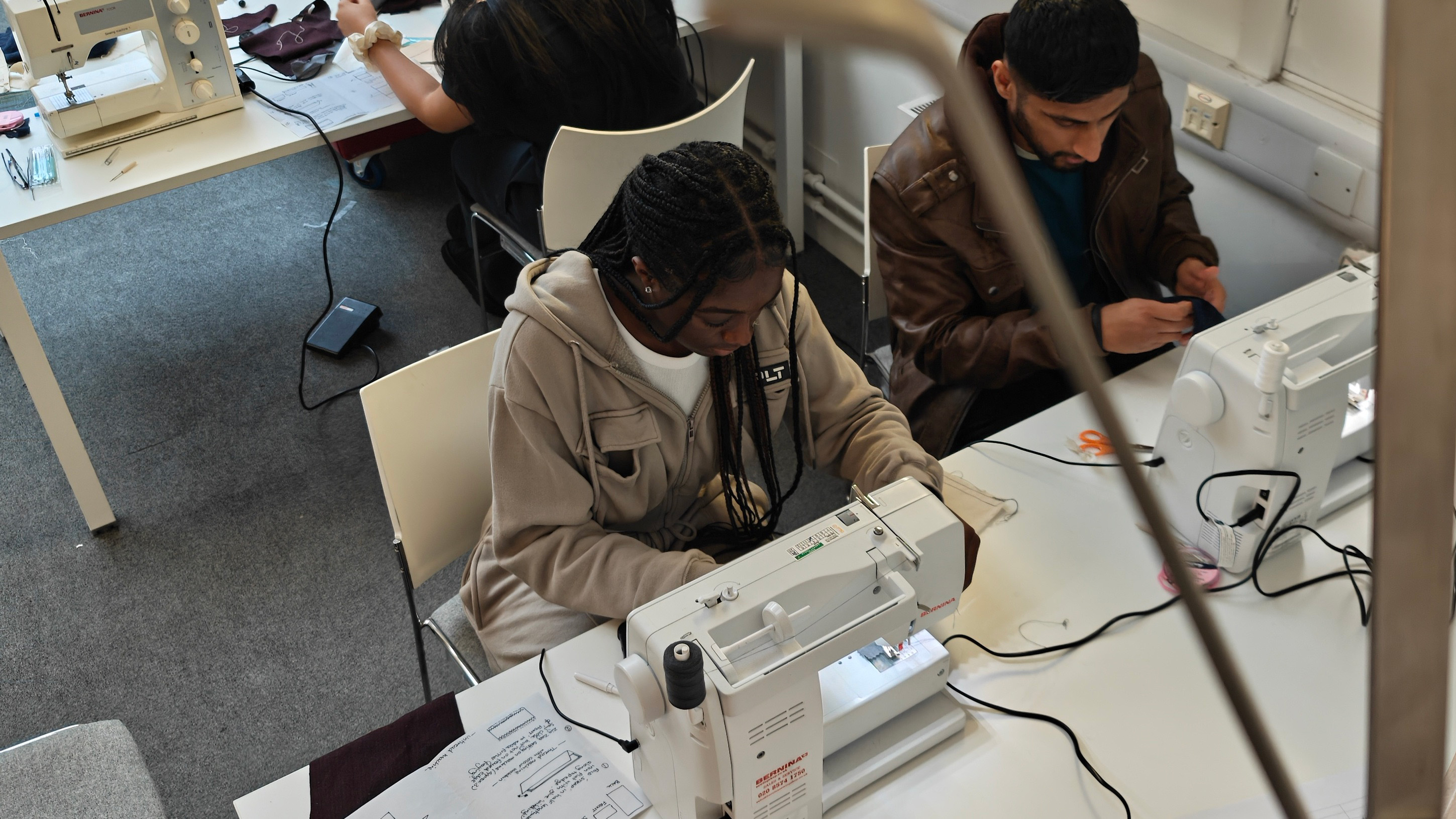 A top-down view of a sewing classroom. A young person with long dark hair, wearing a light hoodie, is sewing at a machine. Another individual in a brown jacket is seated at a neighbouring machine. White tables and sewing materials are visible in the background.