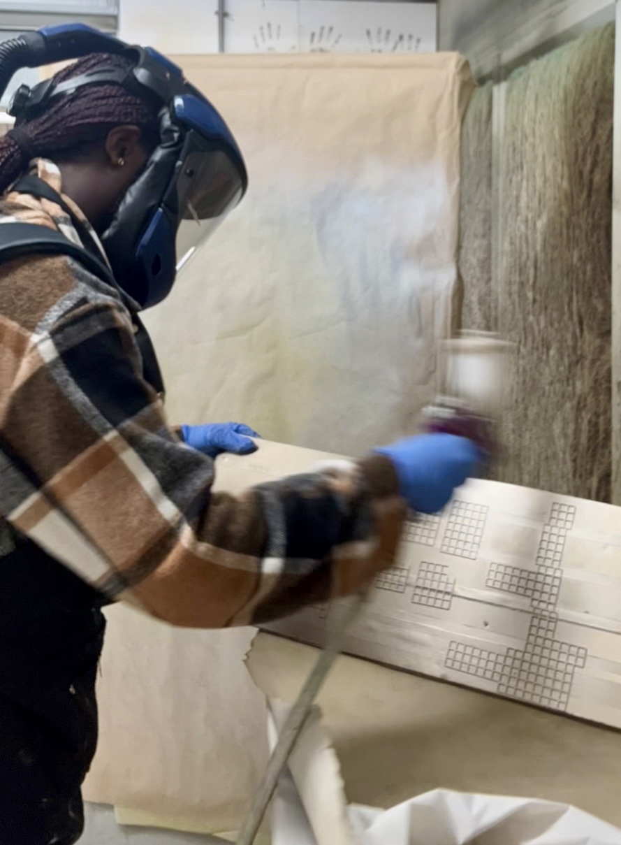 A person wearing a safety mask and gloves is spray painting a wooden board with a grid pattern. The background features brown paper and greenish textures, indicating a workshop environment.