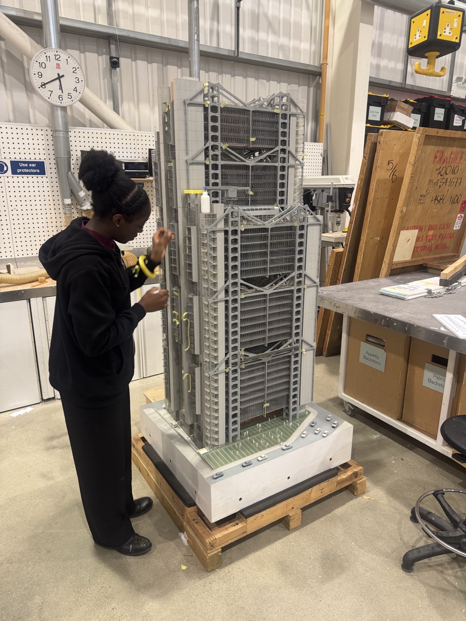 A woman in a black hoodie examines a large architectural model of a building on a wooden platform in a workshop. The model features intricate details and is positioned on a flat surface surrounded by various tools and materials.
