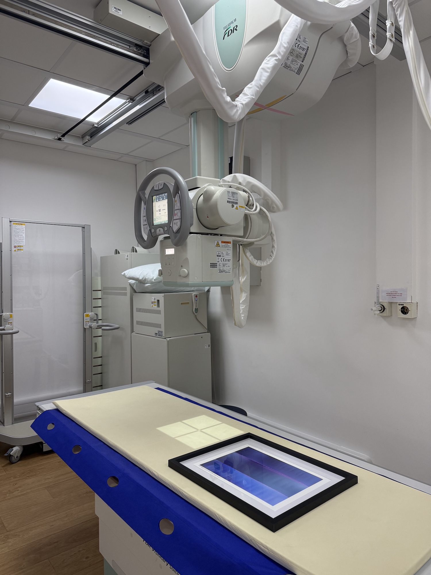 An X-ray machine is positioned above a padded examination table with a blue cover. The background features a medical cabinet and a white wall with visible electrical outlets, indicating a clinical setting.