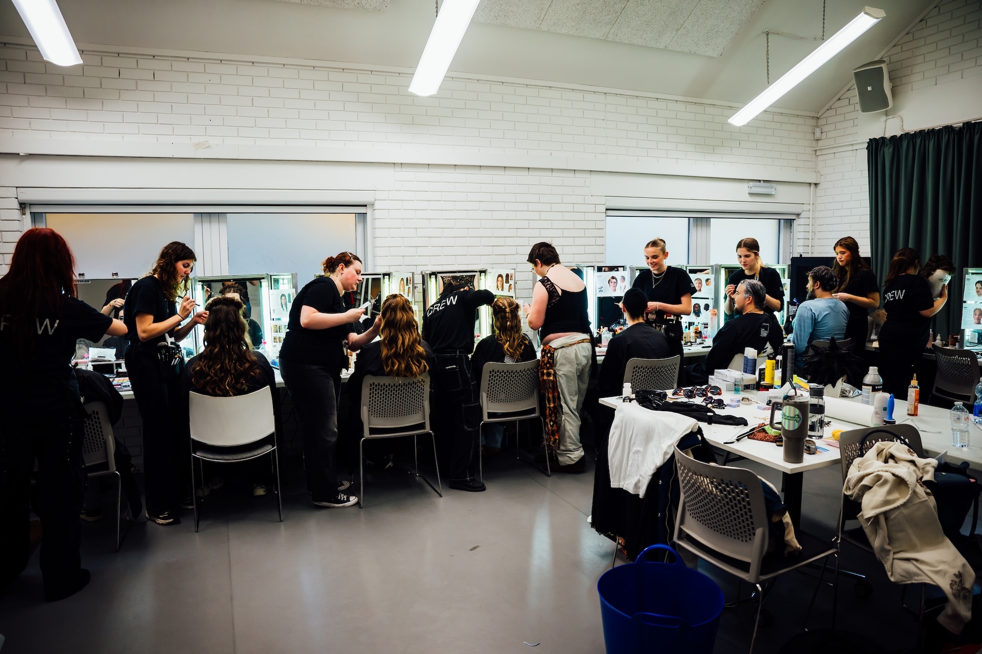 Make-up studio with actors at salon chairs along mirrors, BA (Hons) Make-up, Hair and Prosthetics students artists working with BA (Hons) Acting students, white walls and fluorescent lighting.