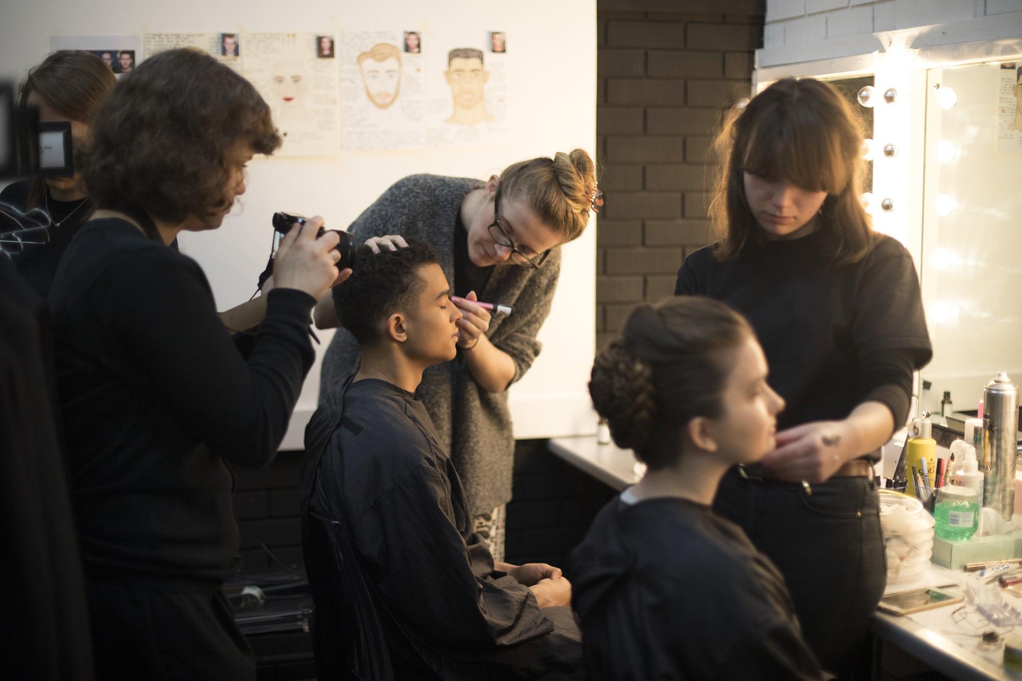 Two people sitting in front of mirrors while make-up artists apply make-up to them. Another person stands to the side and takes a photo.