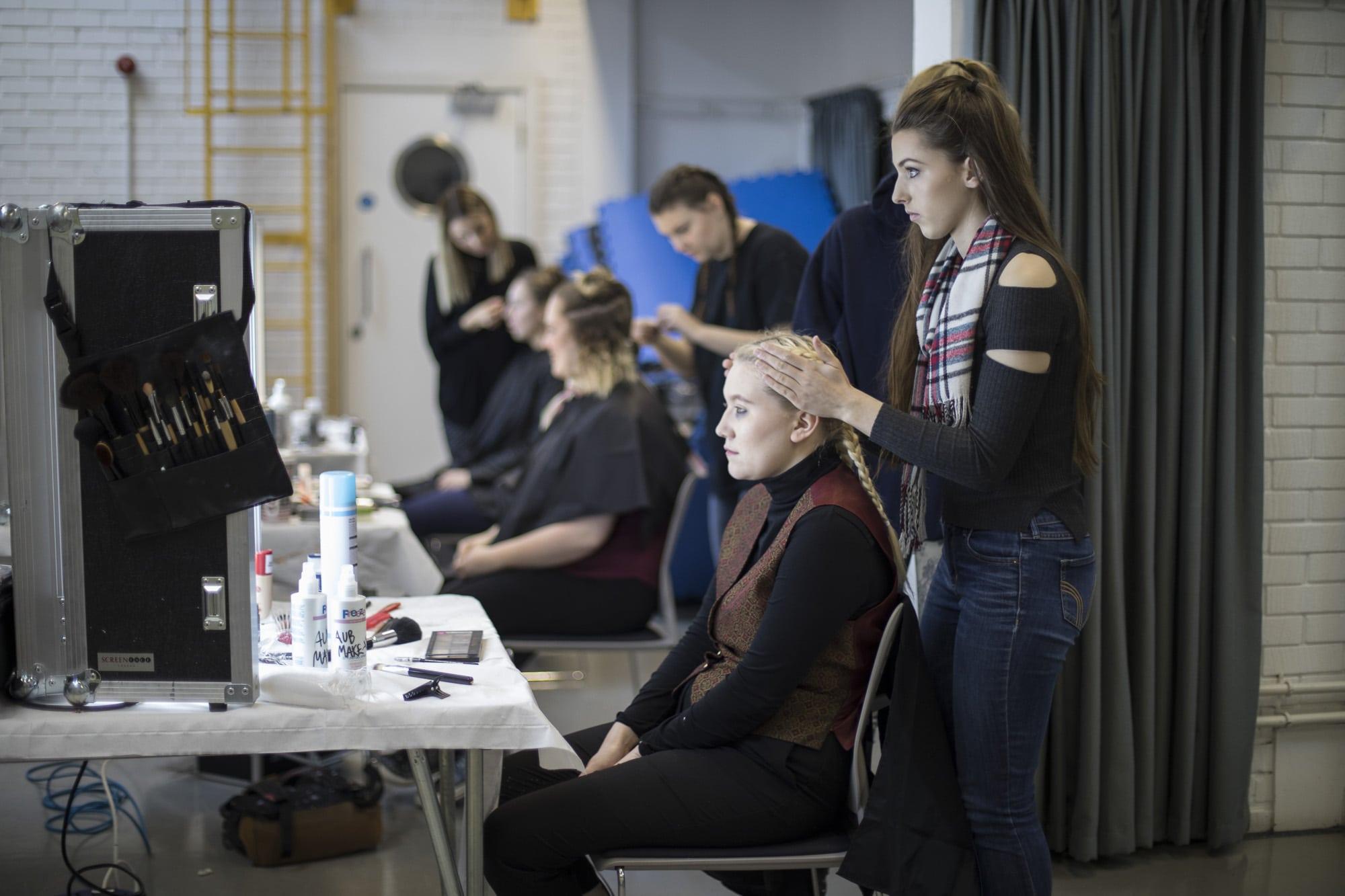 A person sits at a makeup table while another person stands behind them and puts their hands on their hair.
