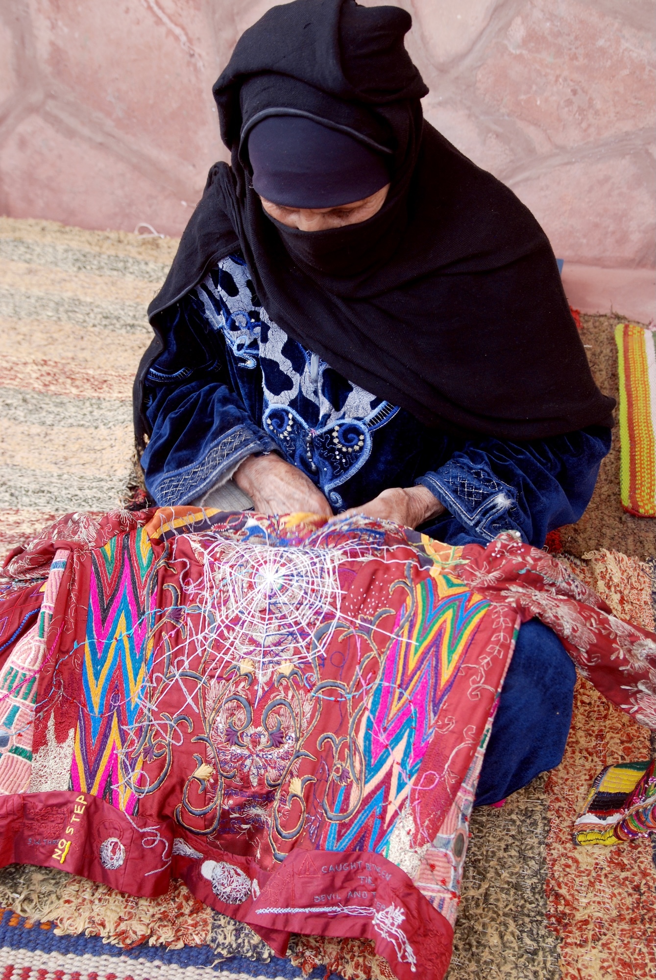 Woman in black robes and headcovering sewing colourful red fabric with zigzag patterns whilst sitting cross-legged on carpet.