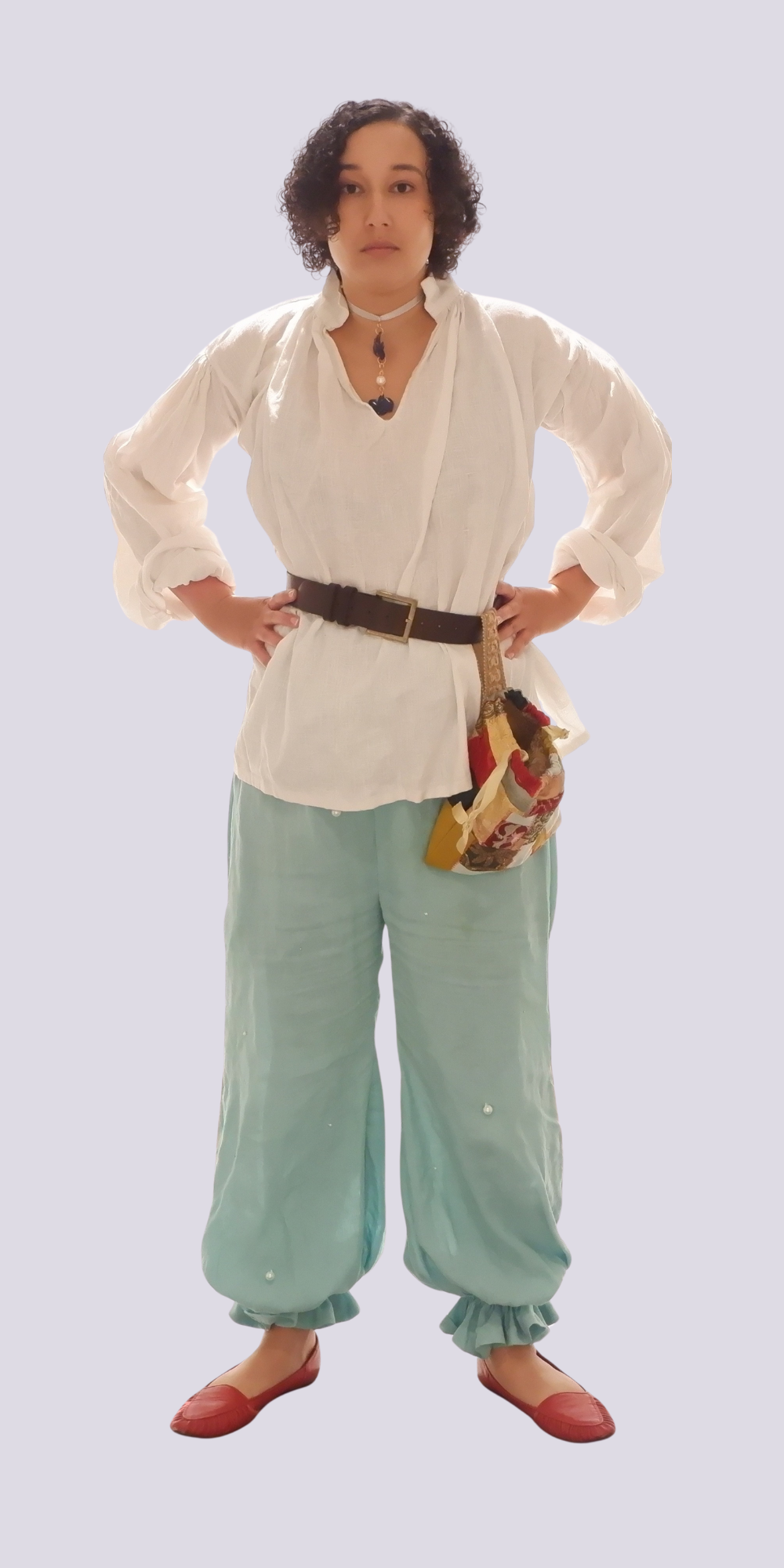 Woman with curly brown hair wearing white shirt, mint green trousers, brown belt with pouch, and red shoes against pale background.