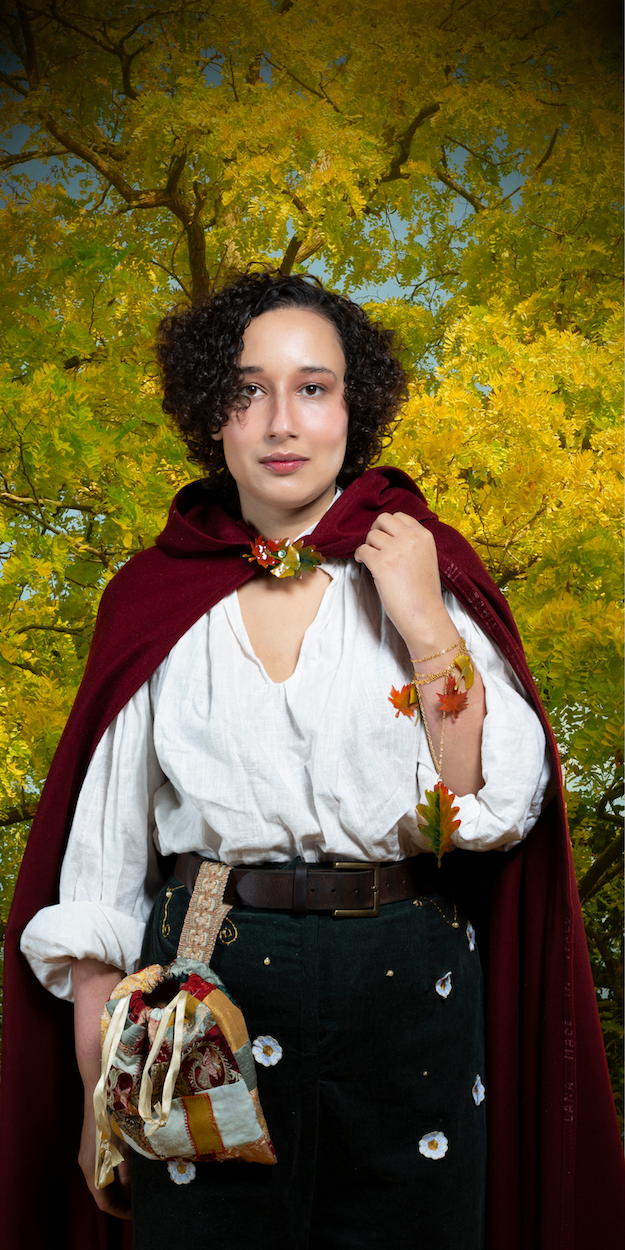 Person with curly dark hair wearing white blouse, dark green skirt, burgundy cloak, holding patterned bag beneath yellow autumn trees.