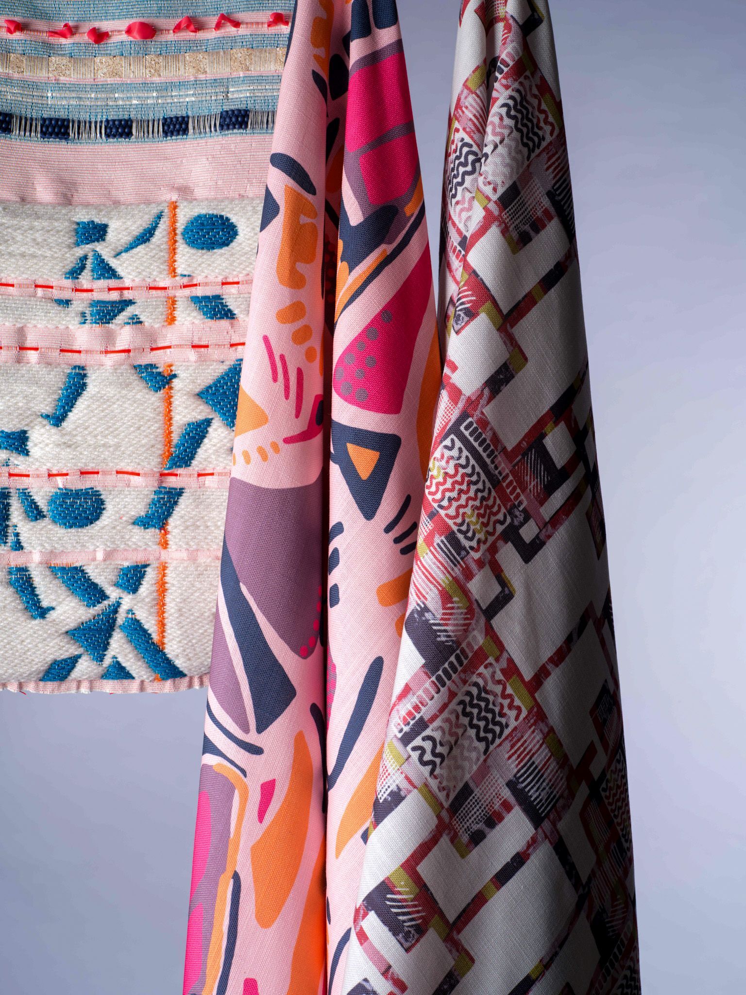 Three patterned textiles hanging vertically with geometric designs in pink, blue, orange, and red colours against white wall.