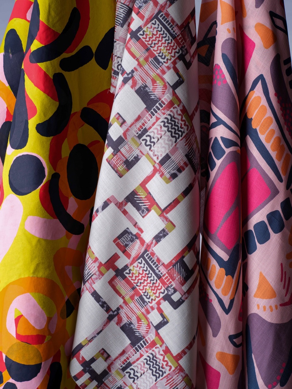 Colourful patterned fabrics with geometric shapes: yellow and red with black curves, white with multicoloured angular forms, pink with navy and orange triangular motifs.
