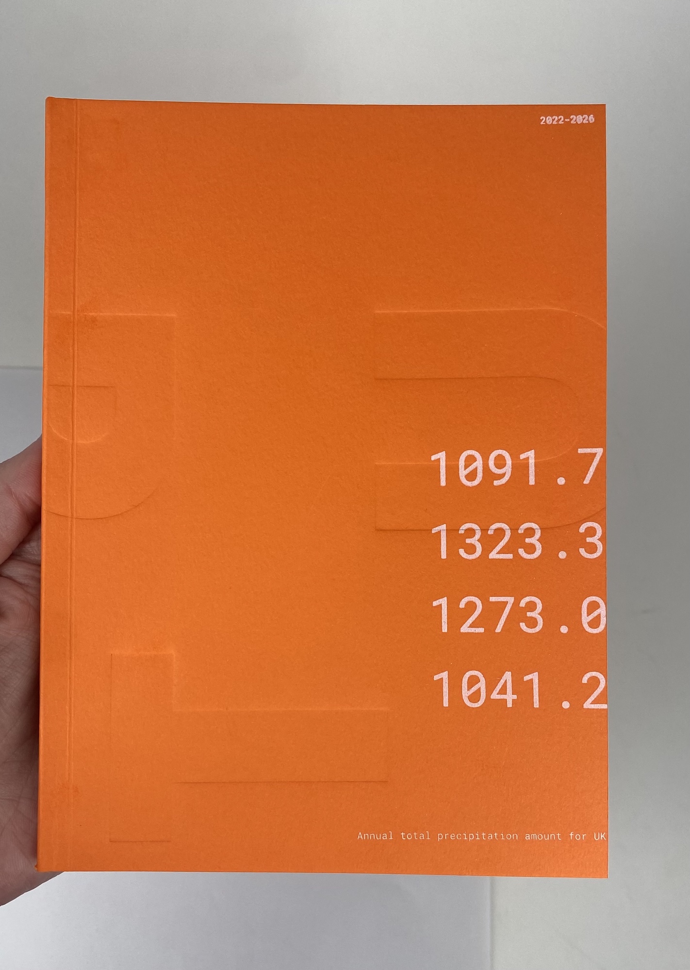 An orange book or report cover with large white numbers on the right side, held by a hand against a light background.