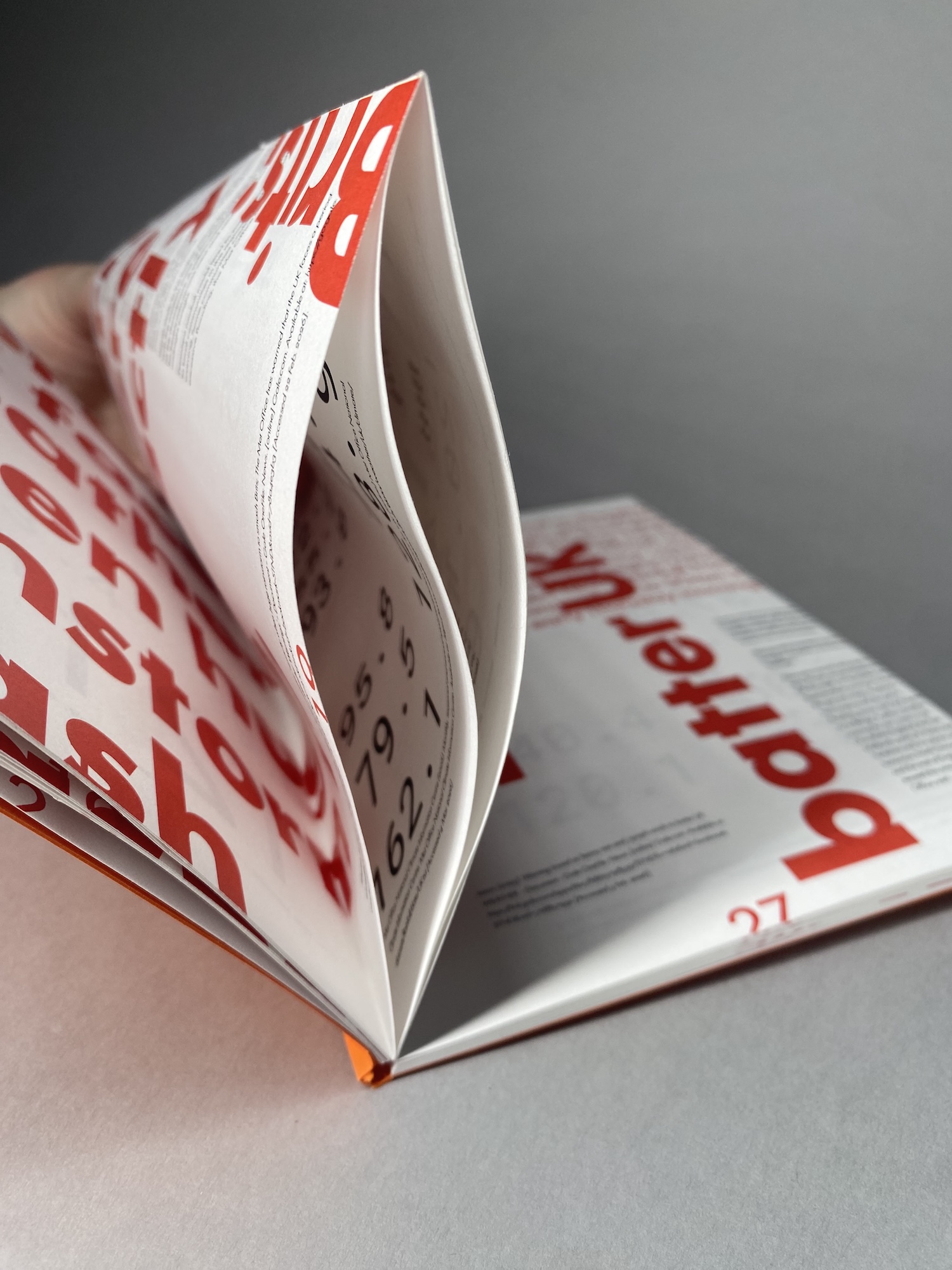 An open magazine with red headline text is standing upright on a grey surface, with another page lying flat behind it.