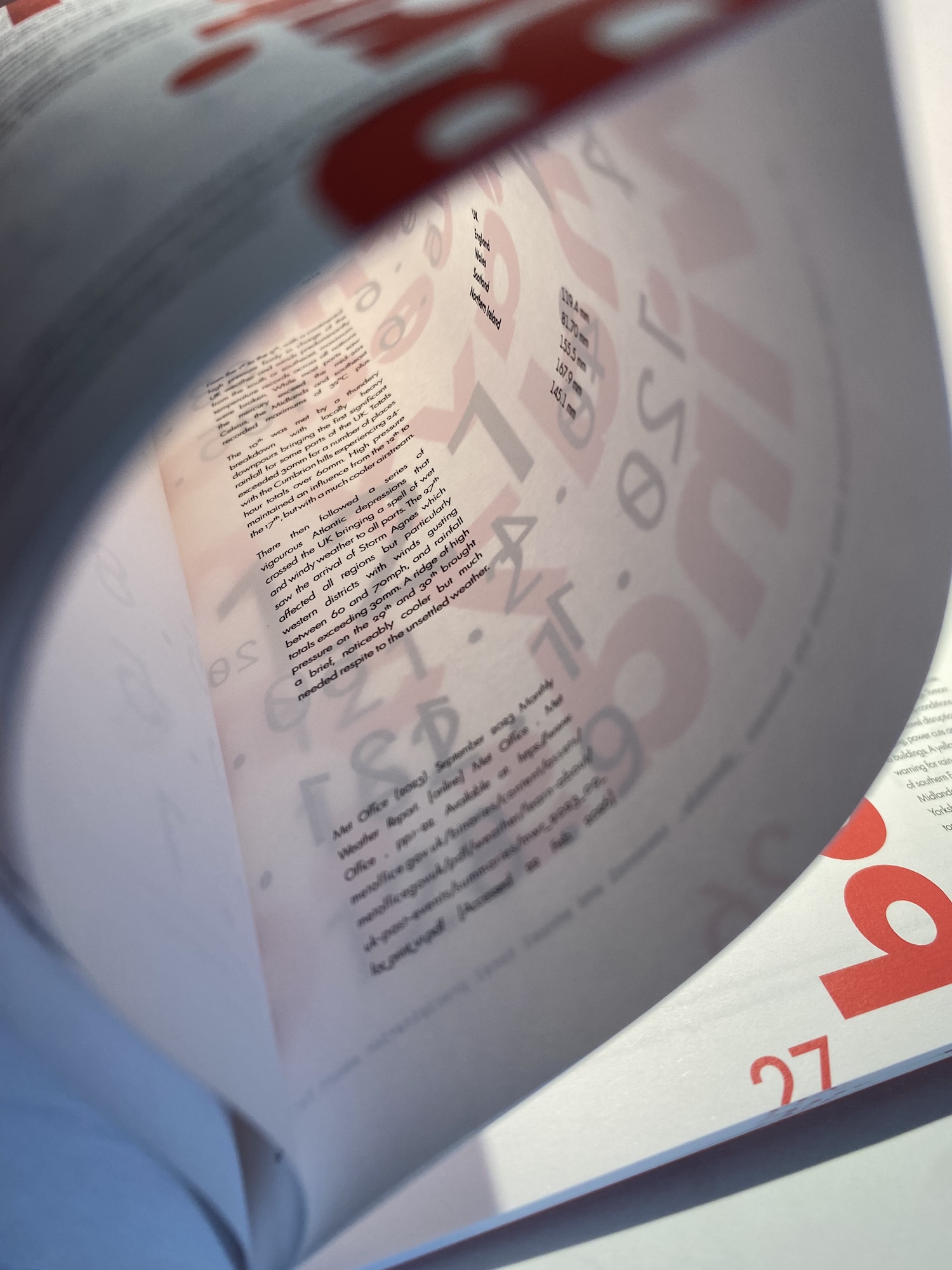 An open magazine page showing blocks of text and large pale pink typography, viewed through a curved page in the foreground.