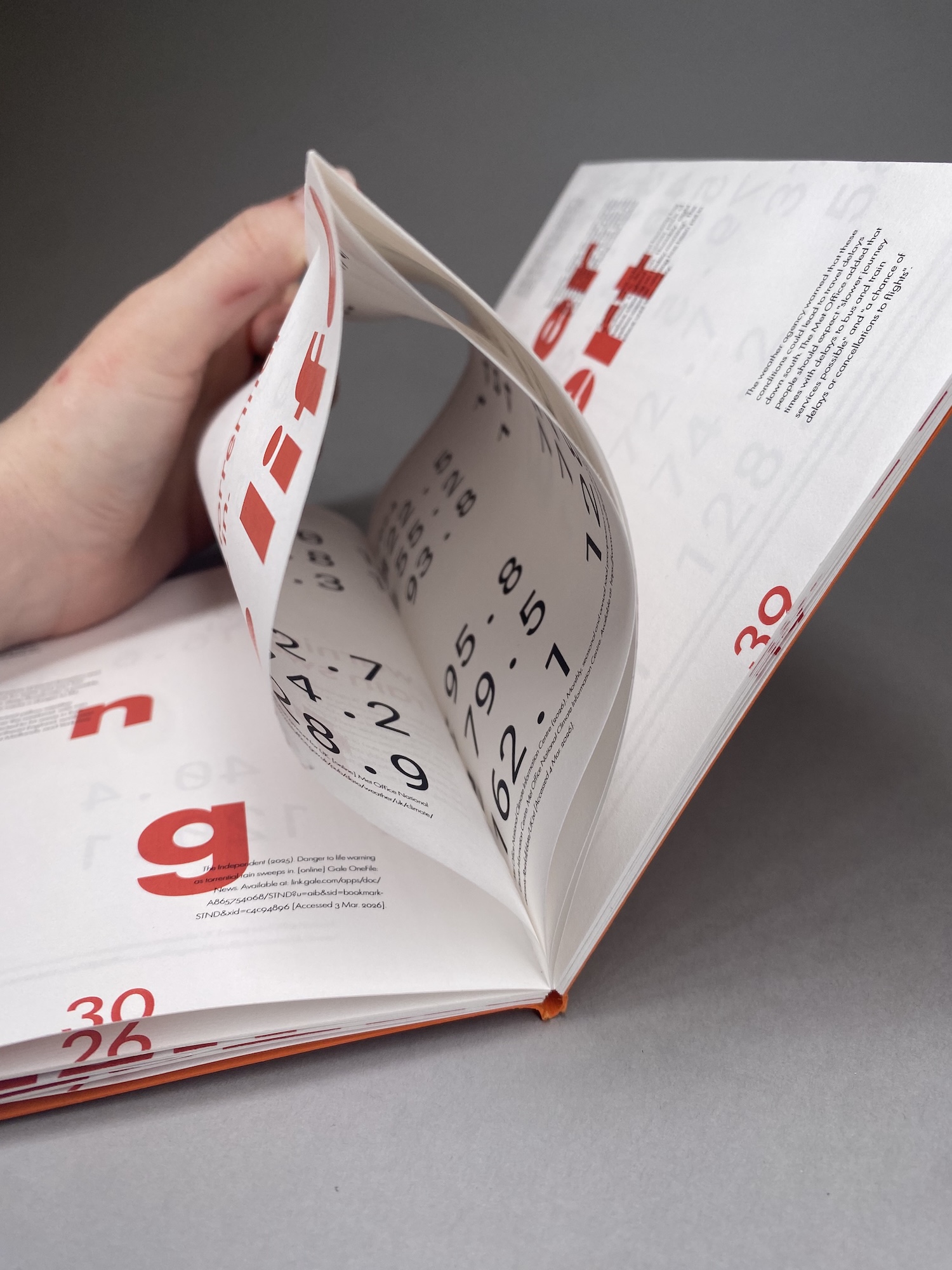 A hand turns the pages of an open calendar or planner on a grey surface, showing large red dates and black numbers.