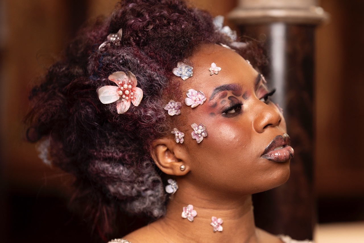 Black woman in profile with elaborate updo hairstyle decorated with pink flowers and crystal gems on her face and neck.
