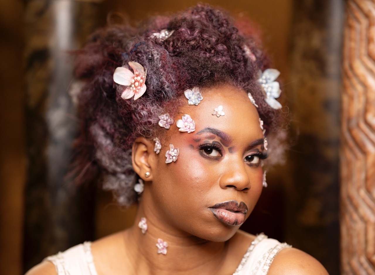 Woman with dark curly hair adorned with pink and white flowers, wearing crystal face gems and pearl earrings.