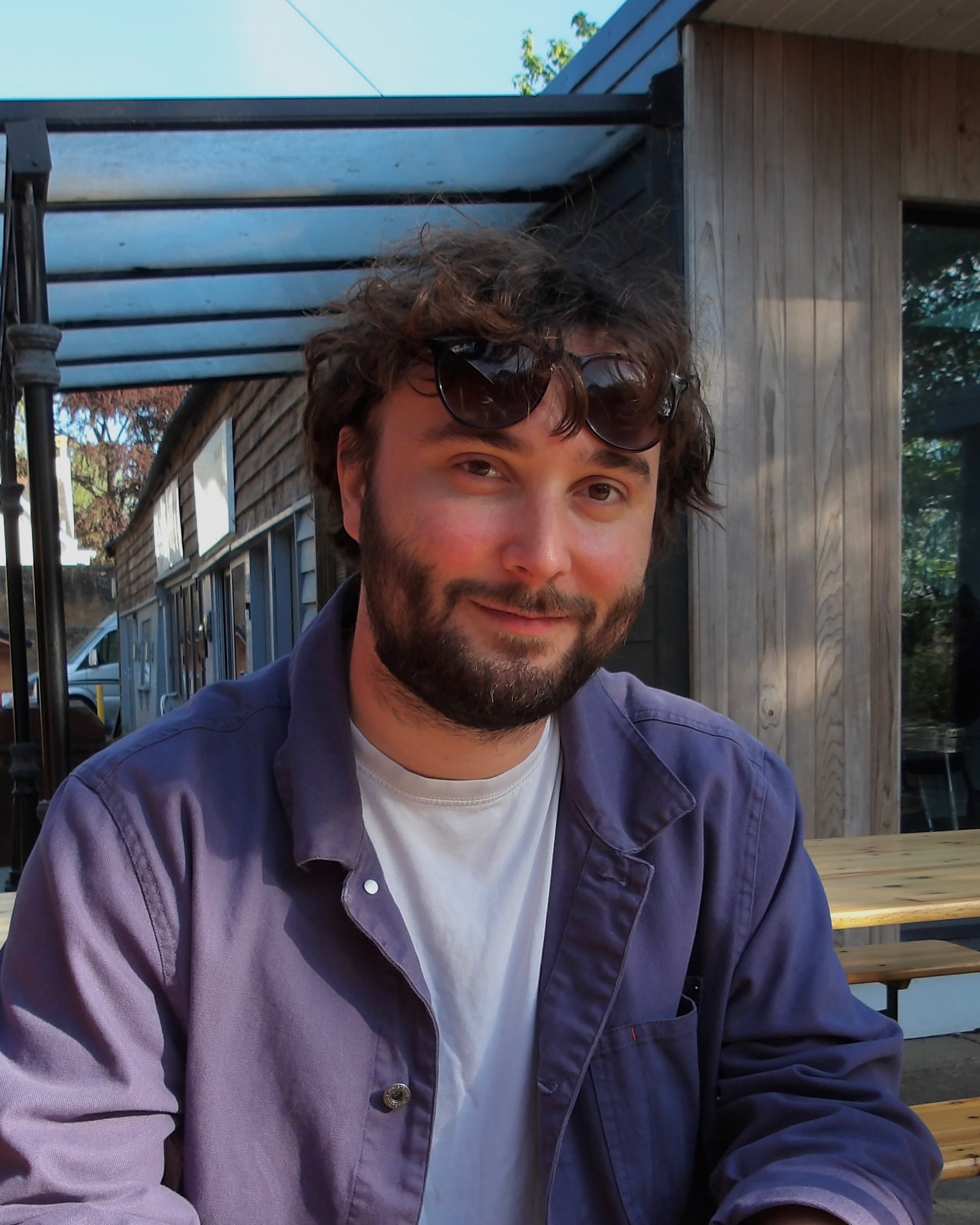 Headshot of Harry Davenport, beard and curly dark hair, sat outside wearing a purple overshirt.