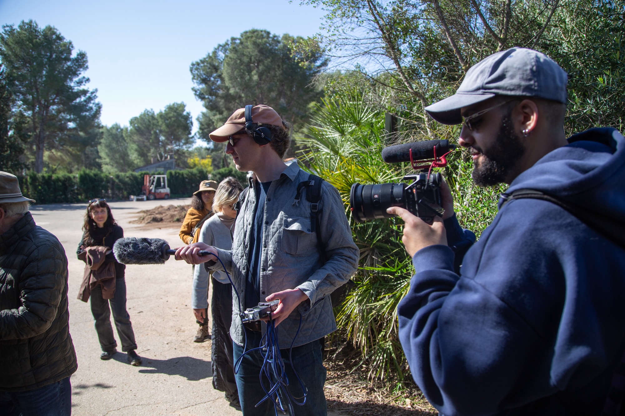 A group of filmmakers working outdoors. One man holds a microphone while wearing headphones, and another man operates a camera. Two women stand nearby, and trees and a vehicle are visible in the background.