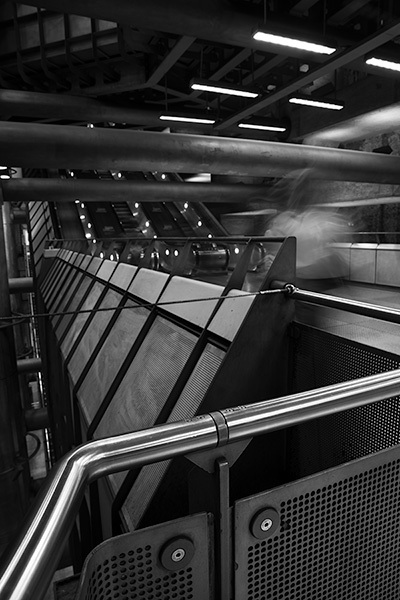Black and white interior showing curved metal handrails, perforated panels, fluorescent ceiling lights, and angular geometric architecture.