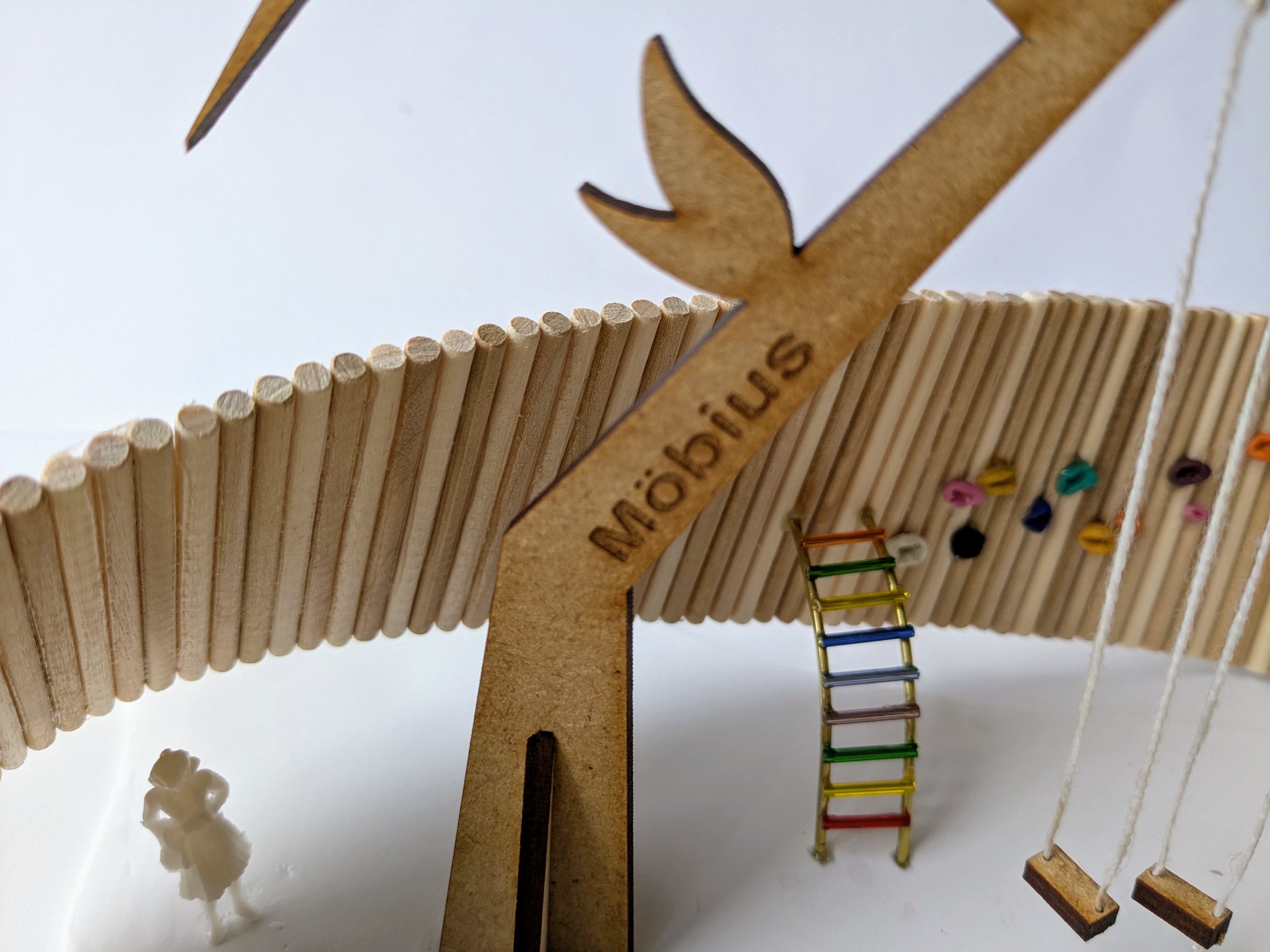 Cardboard model with corrugated blades, small ladder with coloured rungs, and cylindrical base on white surface. Engraved text reads 'Möbius'.