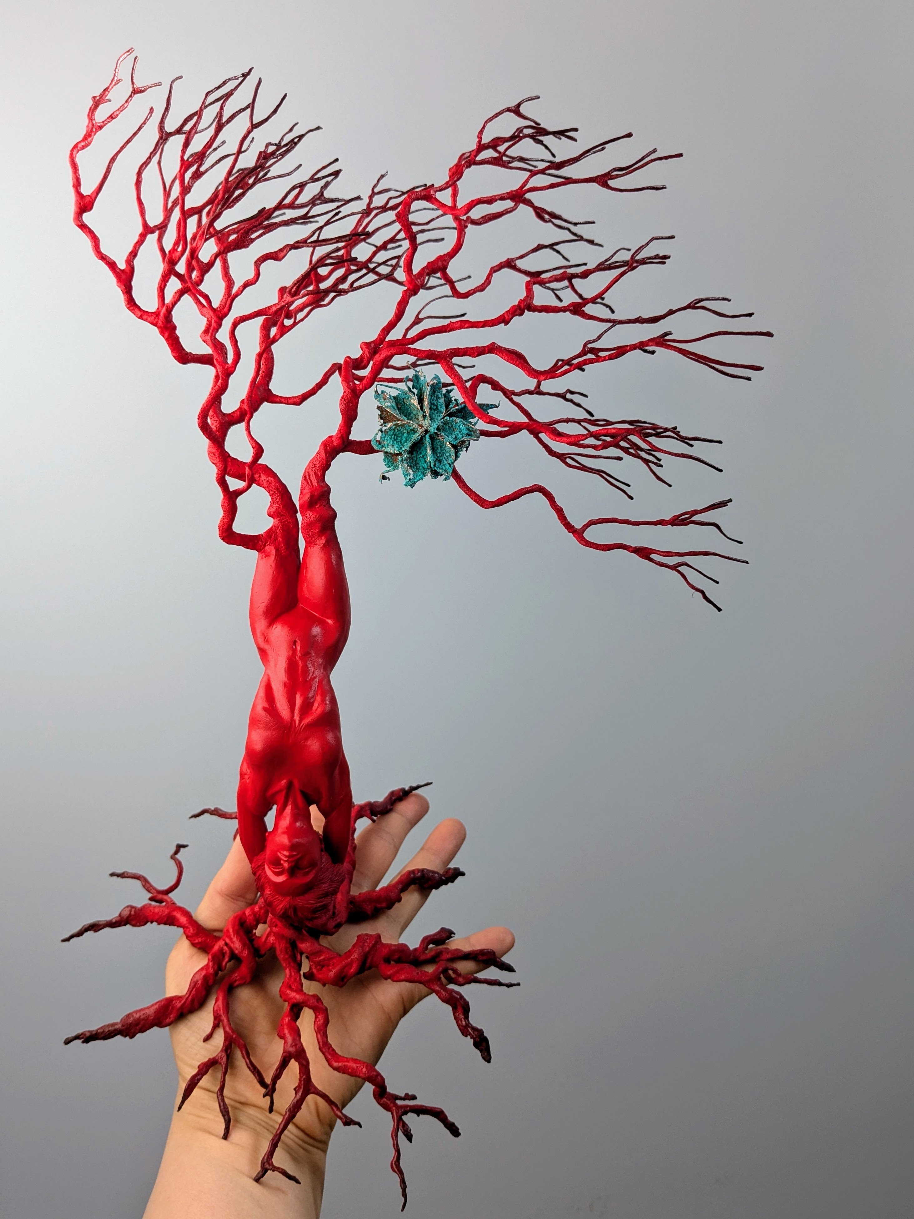 Hand holding red branching tree sculpture with twisted trunk and bare limbs, small green element amongst branches, grey background.