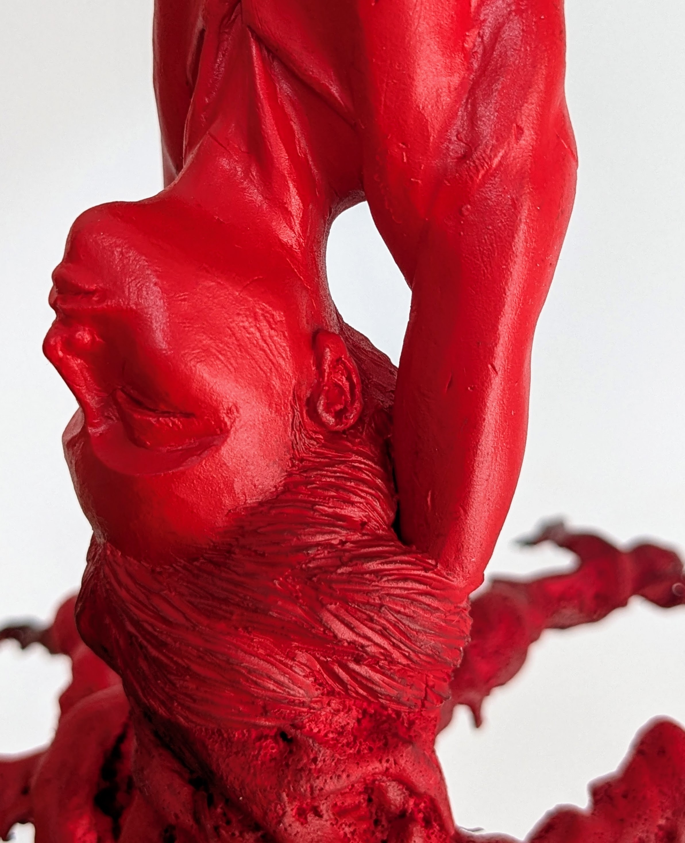 Close-up on red humanoid tree-like sculpture, showing details of upside-down face and hair with closed eyes.