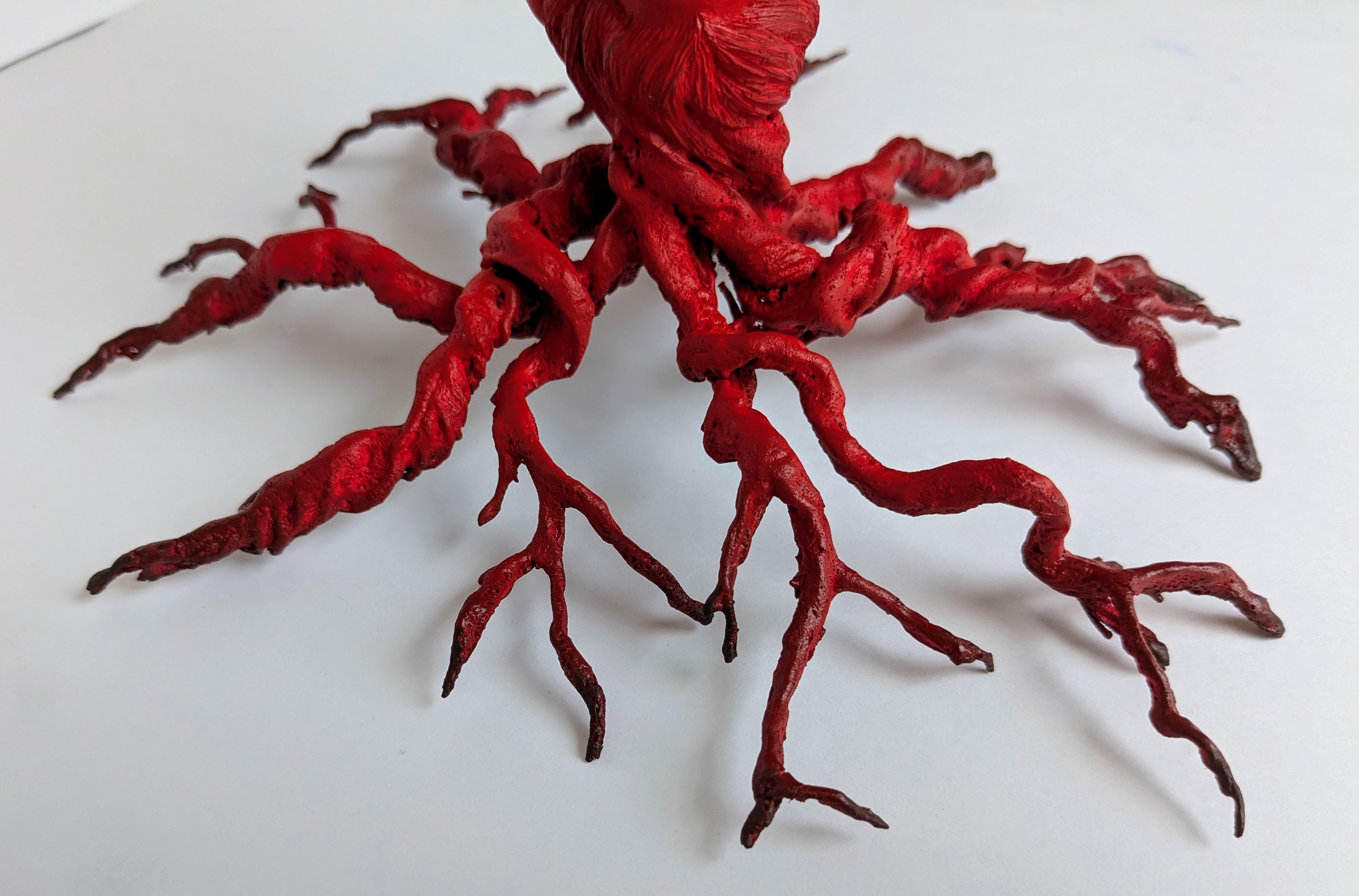 Red sculptural form with multiple twisted, branch-like appendages extending outward from central mass on white surface.
