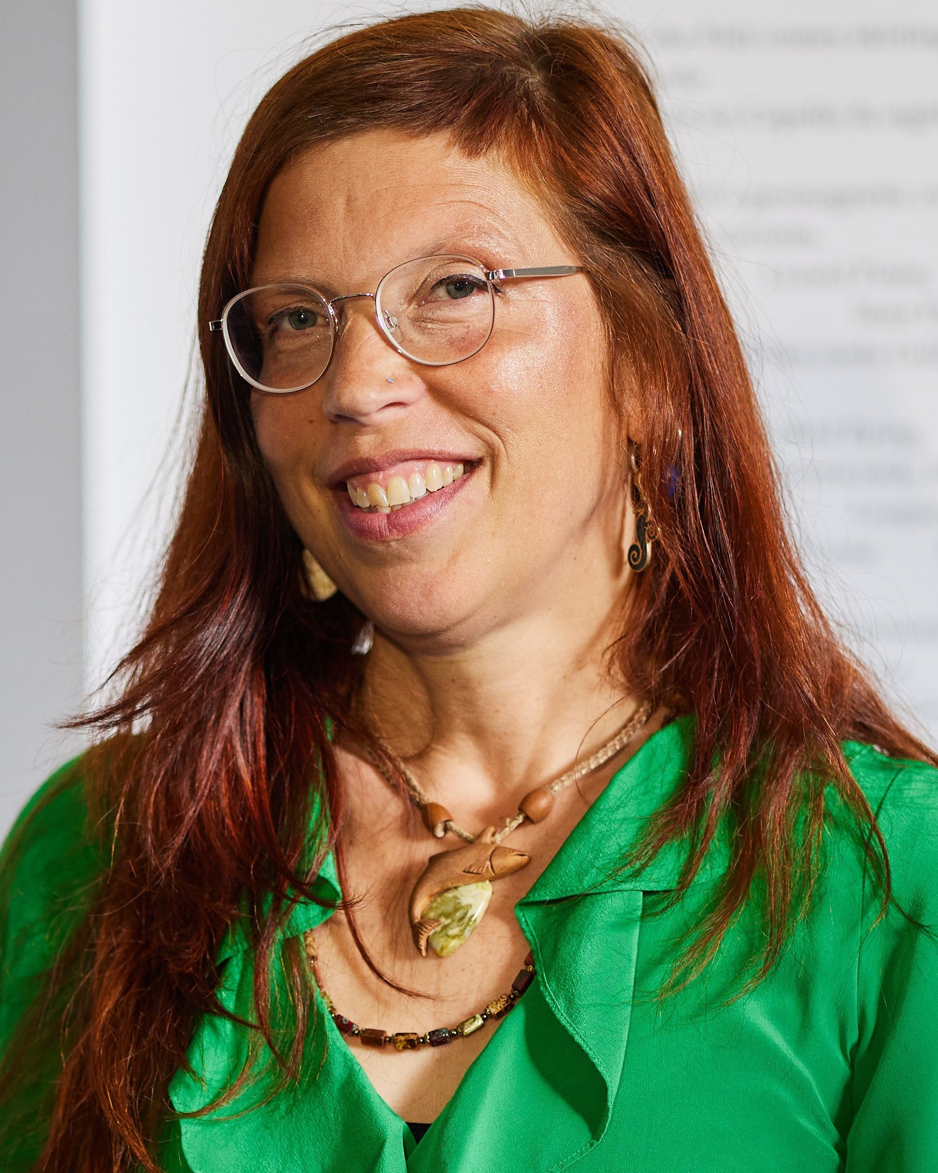 Woman with reddish-brown hair wearing wire-rimmed glasses, green blouse, and gold necklace, smiling at camera.