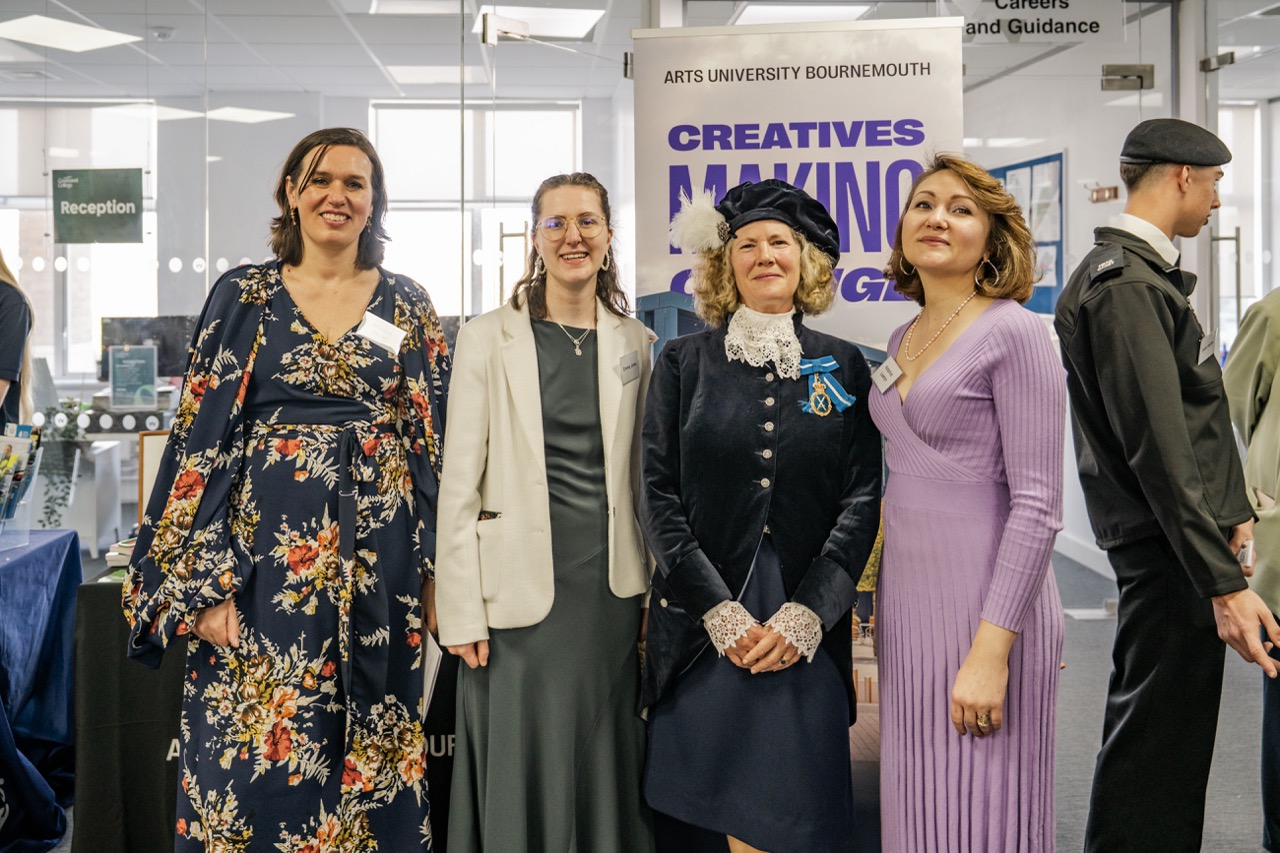 Four women stand together indoors in front of an Arts University Bournemouth Creatives and Making exhibition poster, with glass doors and other people in the background.
