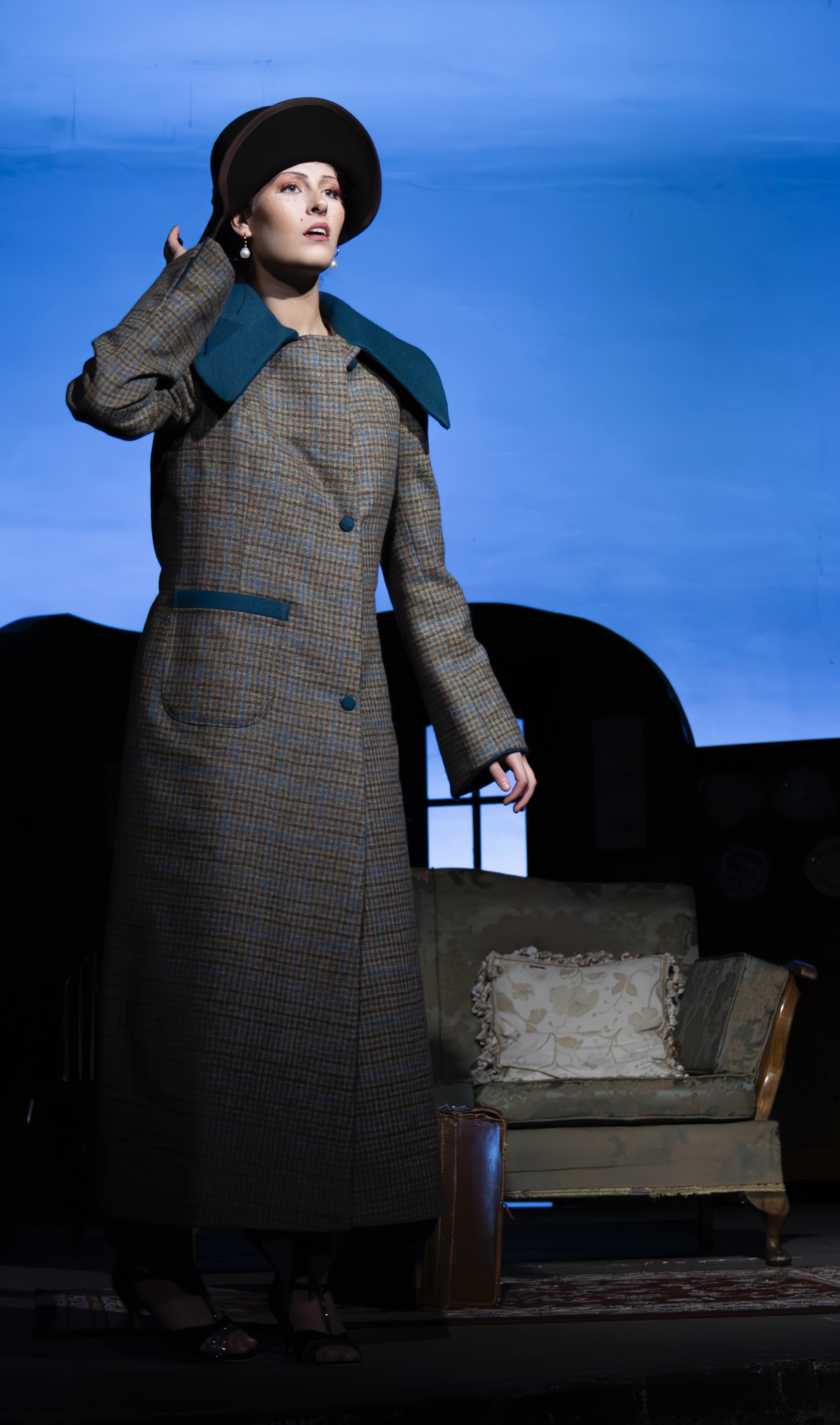 Woman in grey tweed coat with teal trim and black hat stands beside vintage armchair against blue backdrop on theatre stage.