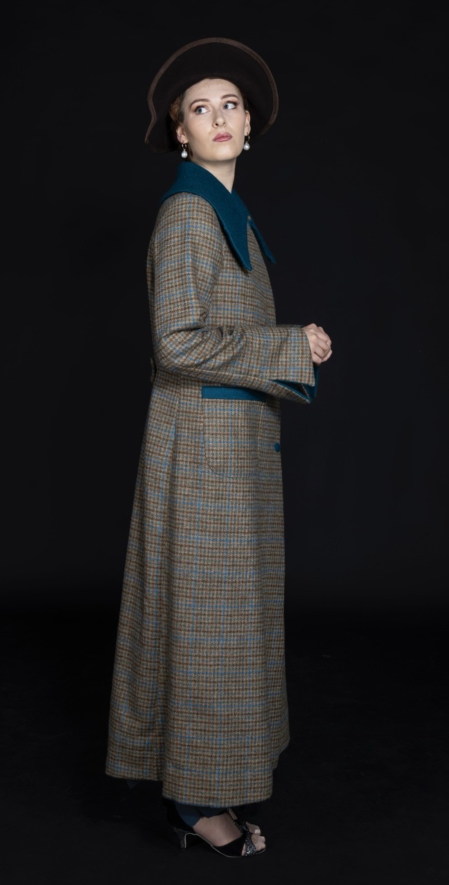 Woman in tweed coat with teal trim and wide-brimmed hat, looking over shoulder against black background.