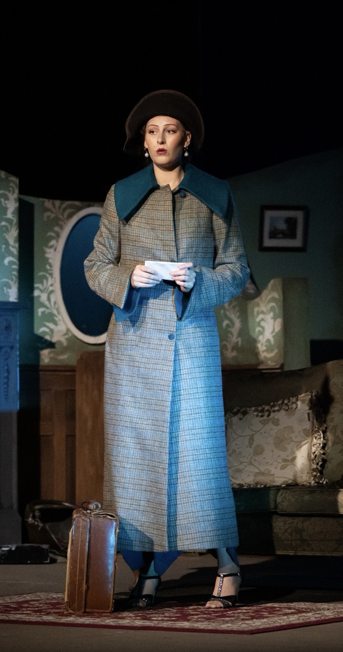 Woman in grey tweed coat with teal collar and hat holding white paper on stage set with patterned wallpaper and vintage furniture.