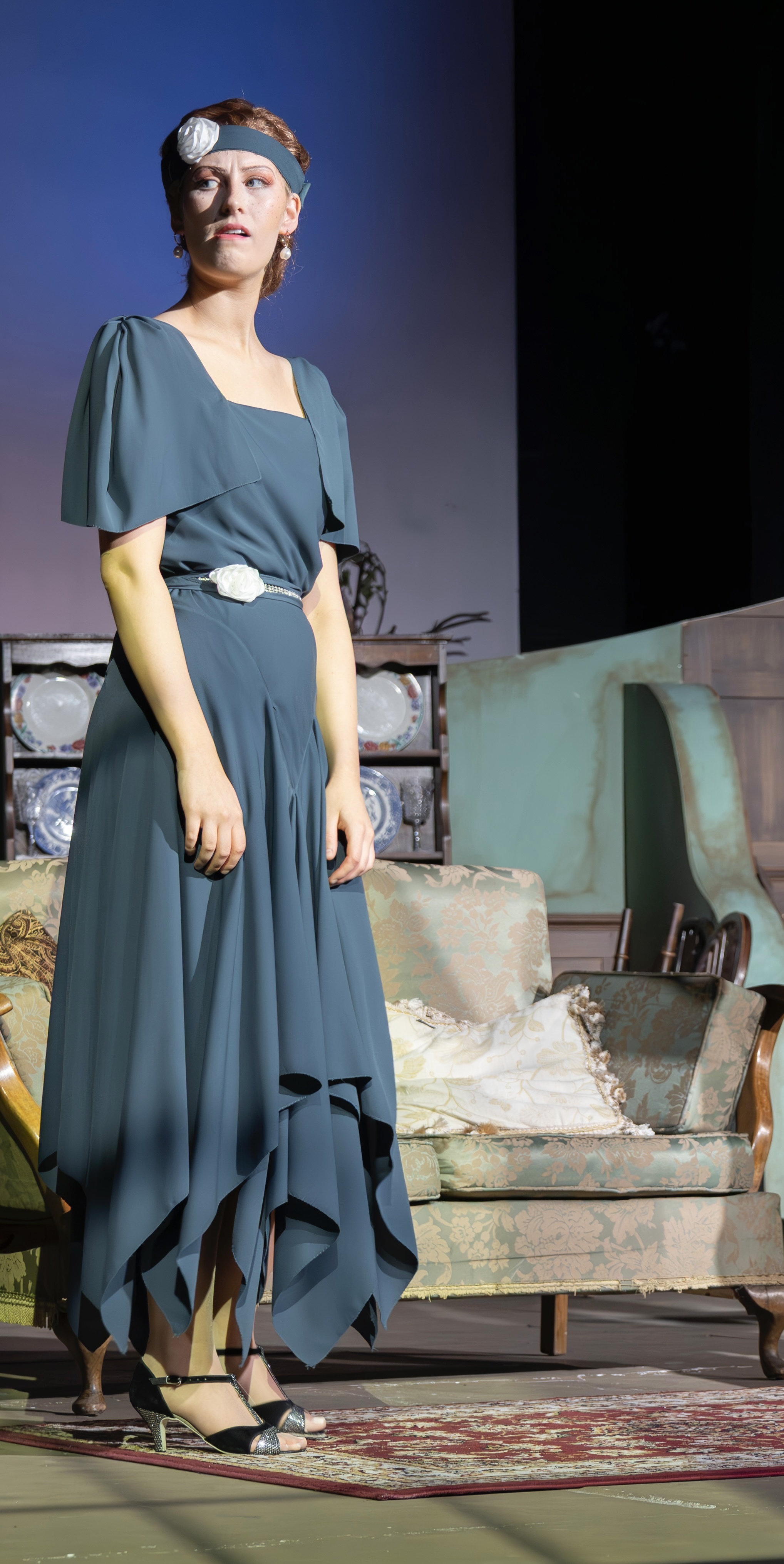 Woman in teal dress with white headband on theatre stage, vintage furniture and rug visible in background.
