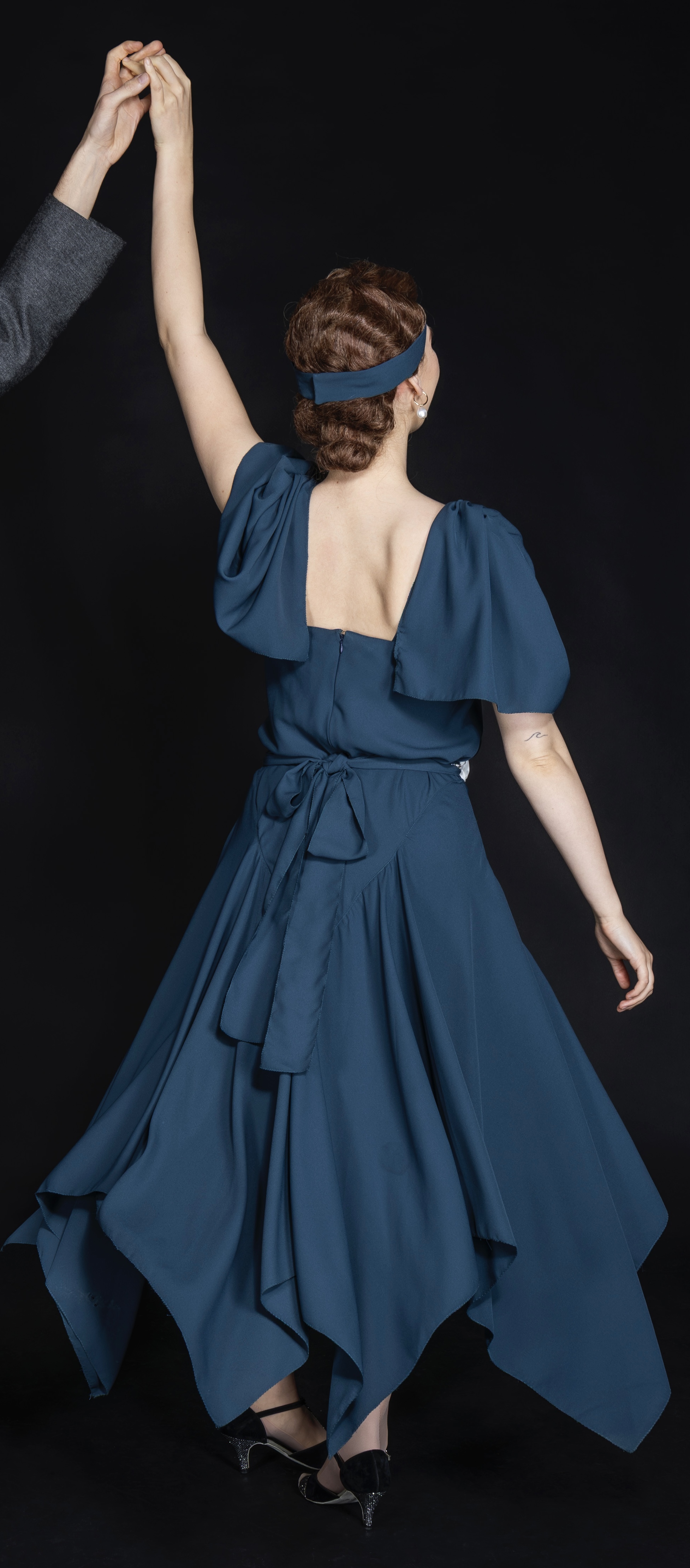 Woman in blue dress with open back and blue headband, viewed from behind with arms raised, against black background.