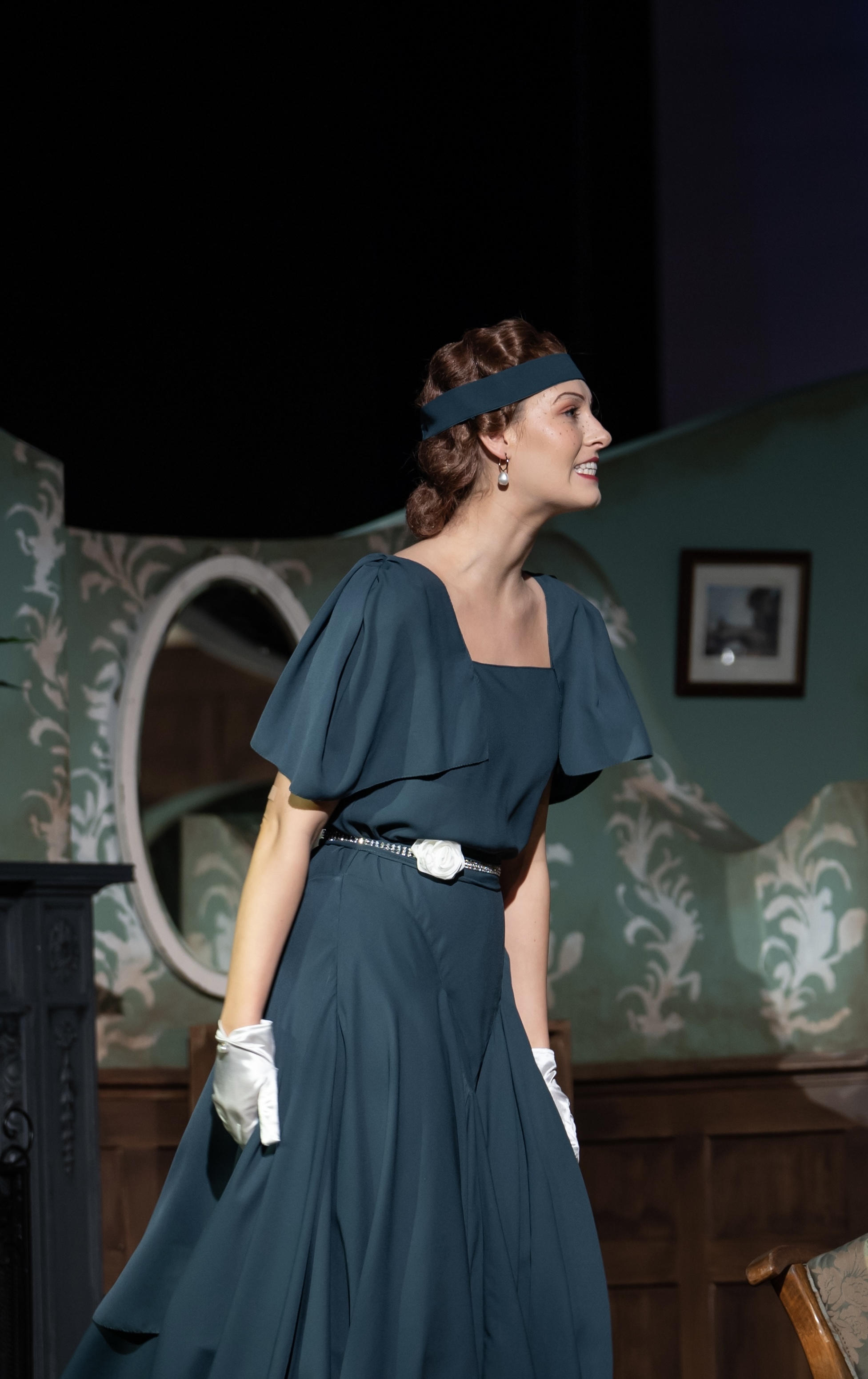Woman in teal 1920s dress with flutter sleeves, headband, and white gloves standing in period room with ornate furniture.